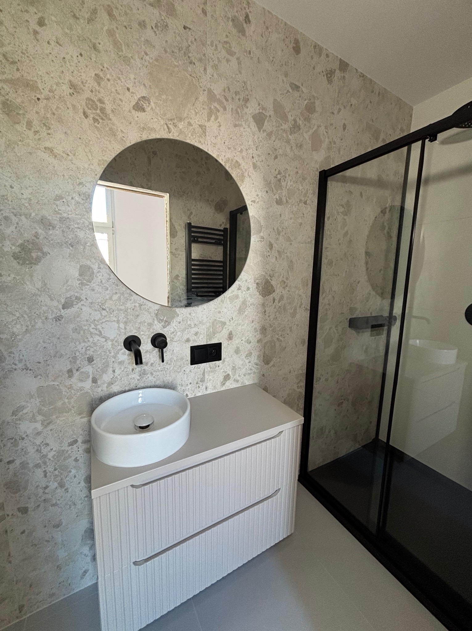 Nowoczesna łazienka z okrągłym lustrem, białą umywalką nablatową na szafce z frezowanymi frontami i kabiną prysznicową z czarnymi profilami na tle marmurowych płytek.