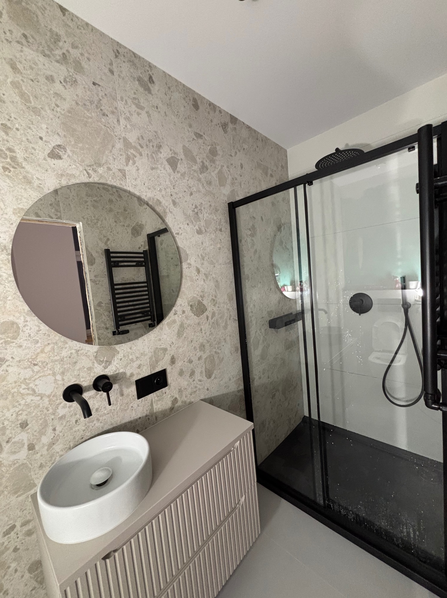 Nowoczesna łazienka z beżowymi płytkami imitującymi kamień, okrągłym lustrem, białą umywalką nablatową i czarną kabiną prysznicową typu walk-in.
