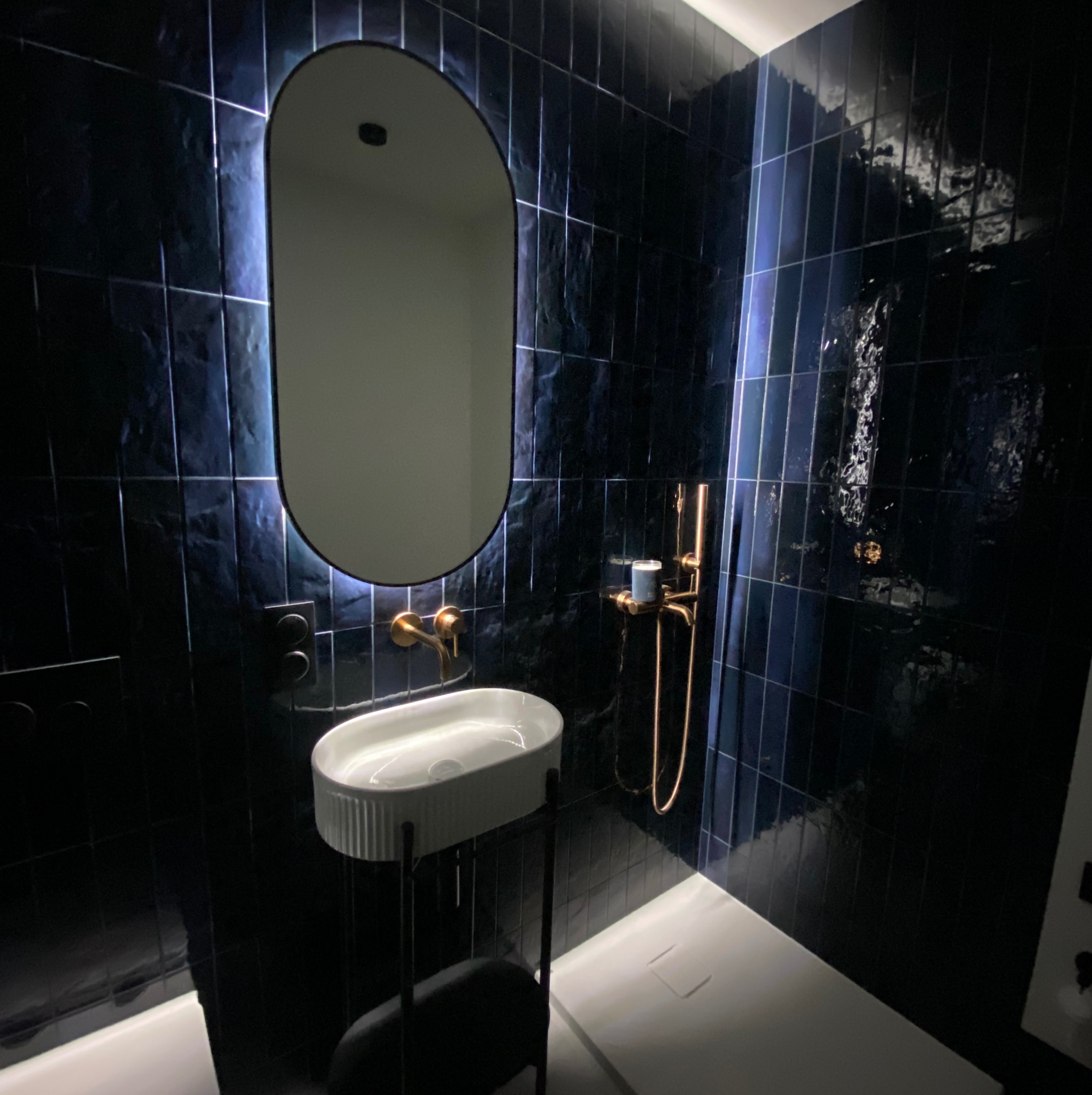 Nowoczesna łazienka z ciemnymi, błyszczącymi płytkami, owalnym lustrem z podświetleniem LED i złotą armaturą. Minimalistyczna umywalka na stelażu.