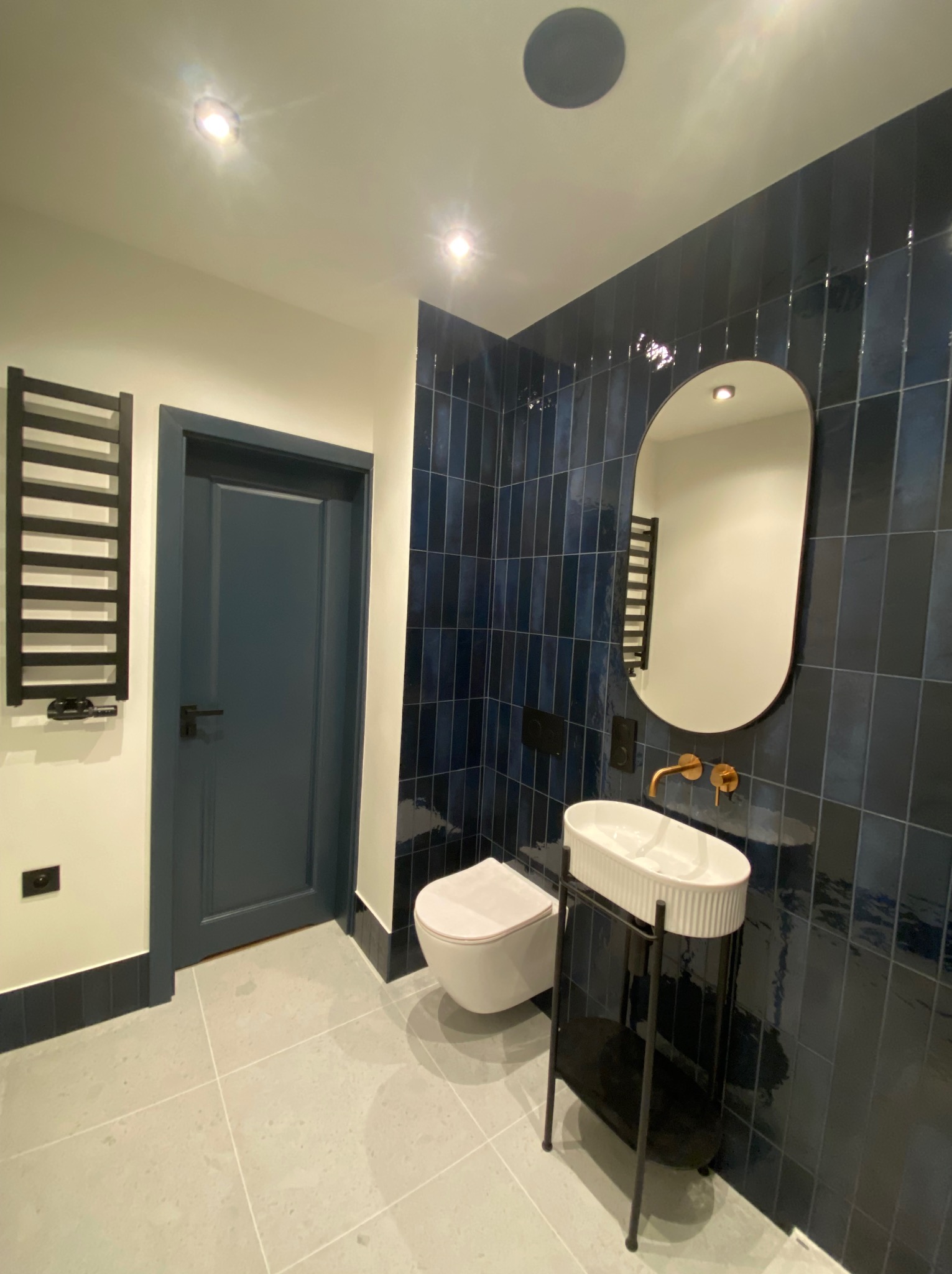 Nowoczesna łazienka z ciemnymi, błyszczącymi płytkami, owalnym lustrem i białą umywalką na czarnym stelażu. Widoczny grzejnik drabinkowy i drzwi w kolorze płytek.