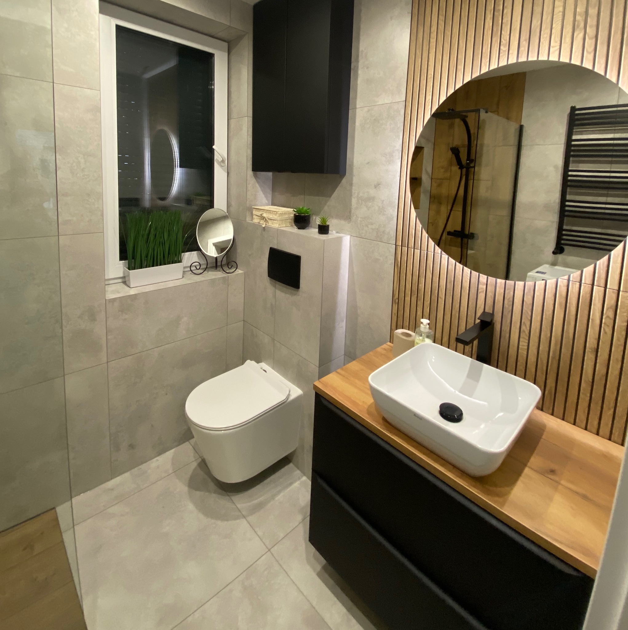 Nowoczesna łazienka z drewnianymi lamelami, okrągłym lustrem i białą umywalką nablatową. Minimalistyczny design, szare płytki i czarne akcenty.