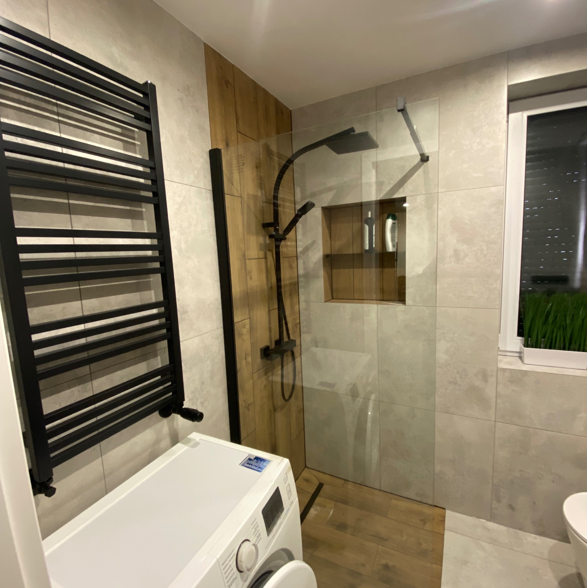 Nowoczesna łazienka po remoncie: kabina walk-in z czarną armaturą, grzejnik drabinkowy, pralka. Płytki imitujące drewno i kamień. Minimalistyczny design.