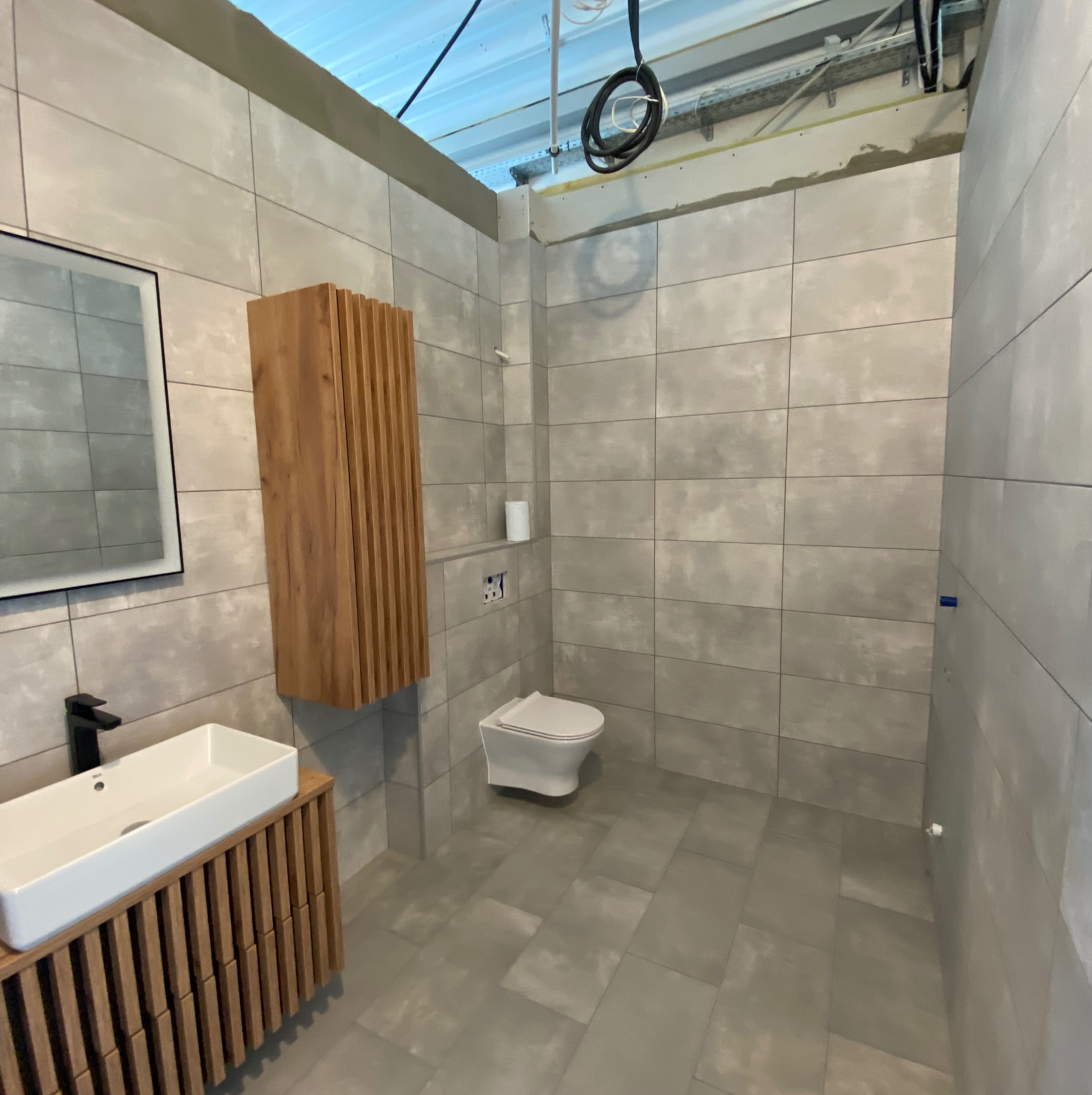 Wykończona łazienka w szarościach z drewnianymi akcentami: szafka pod umywalką i wisząca szafka z lamelami. Widoczna toaleta i lustro z podświetleniem.