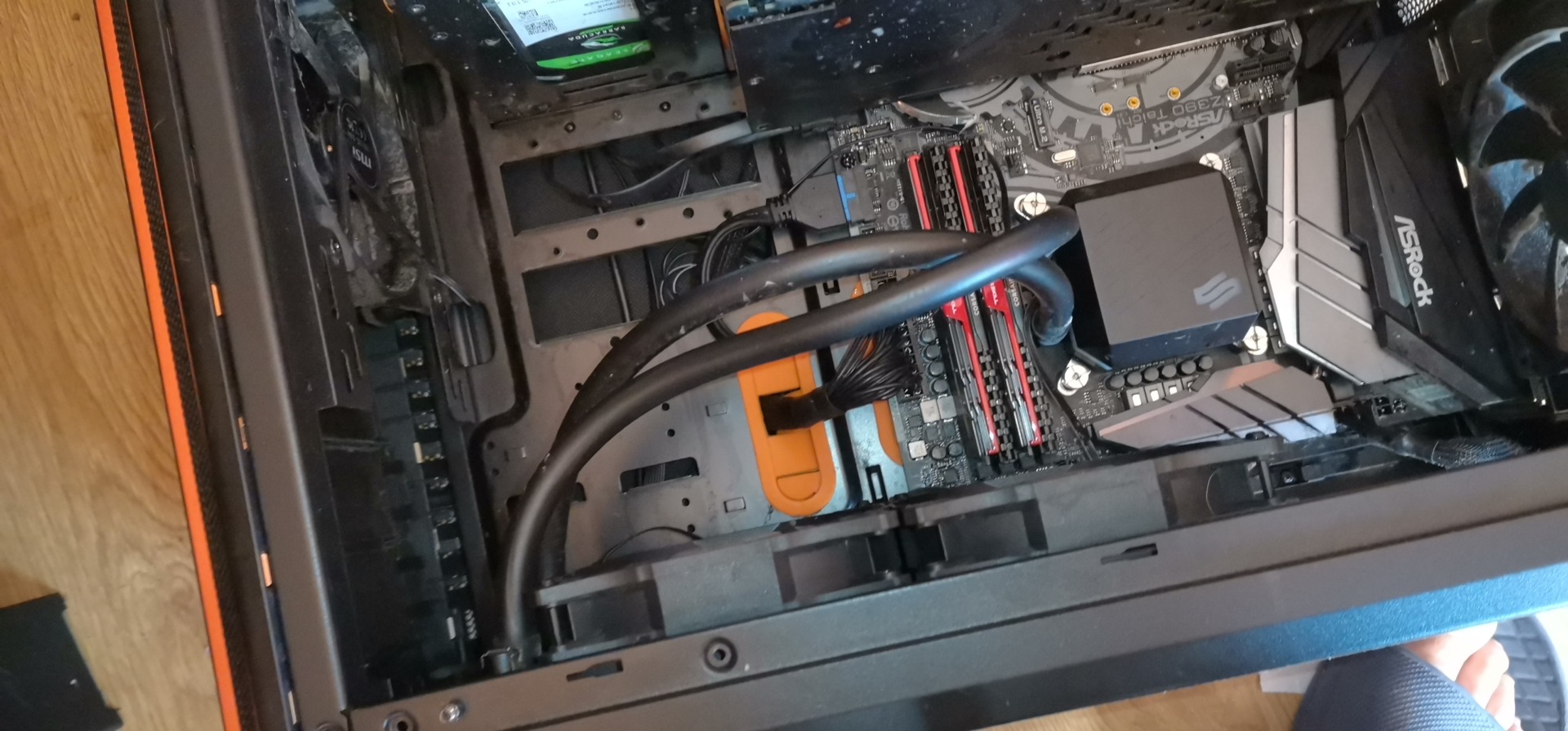 Wnętrze obudowy komputera stacjonarnego z widoczną płytą główną ASRock, chłodzeniem wodnym i pamięcią RAM Corsair. Widoczne okablowanie i elementy montażowe.