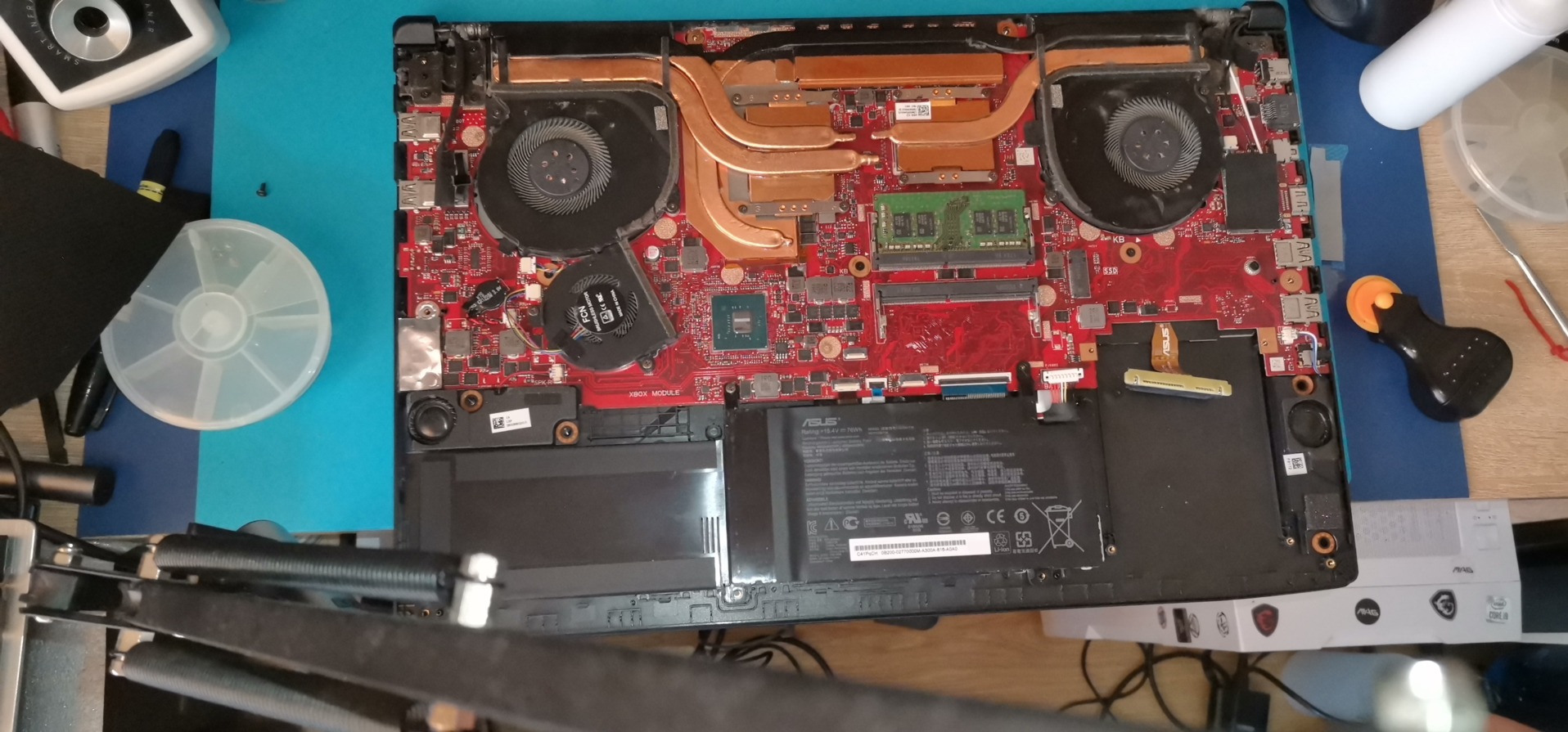 Otwarty laptop ASUS z widoczną płytą główną, układem chłodzenia i baterią. Widoczne narzędzia serwisowe. Układ elementów wskazuje na etap naprawy lub diagnostyki.