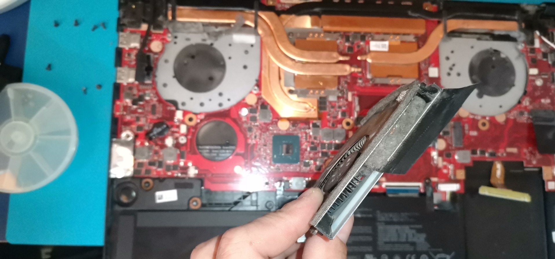 Otwarty laptop z widoczną płytą główną i systemem chłodzenia. W dłoni trzymany wentylator z radiatorem. Widoczne śrubki i narzędzia. Detal naprawy komputera.