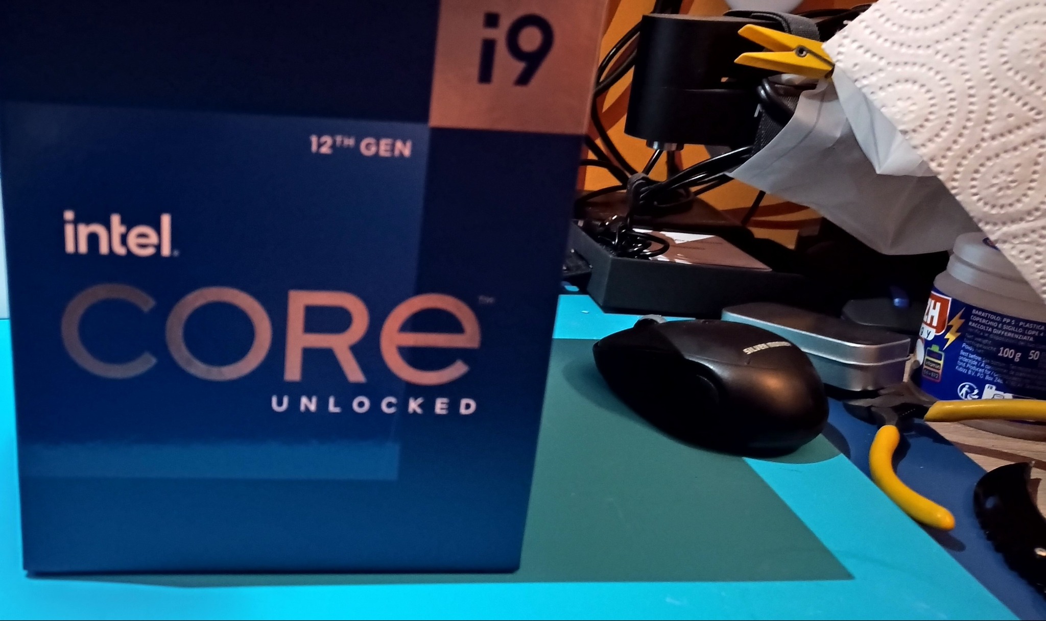 Pudełko procesora Intel Core i9 12th Gen na biurku z myszką, narzędziami i akcesoriami. Widoczne logo Intel i napis CORE UNLOCKED. Niebieska kolorystyka pudełka.