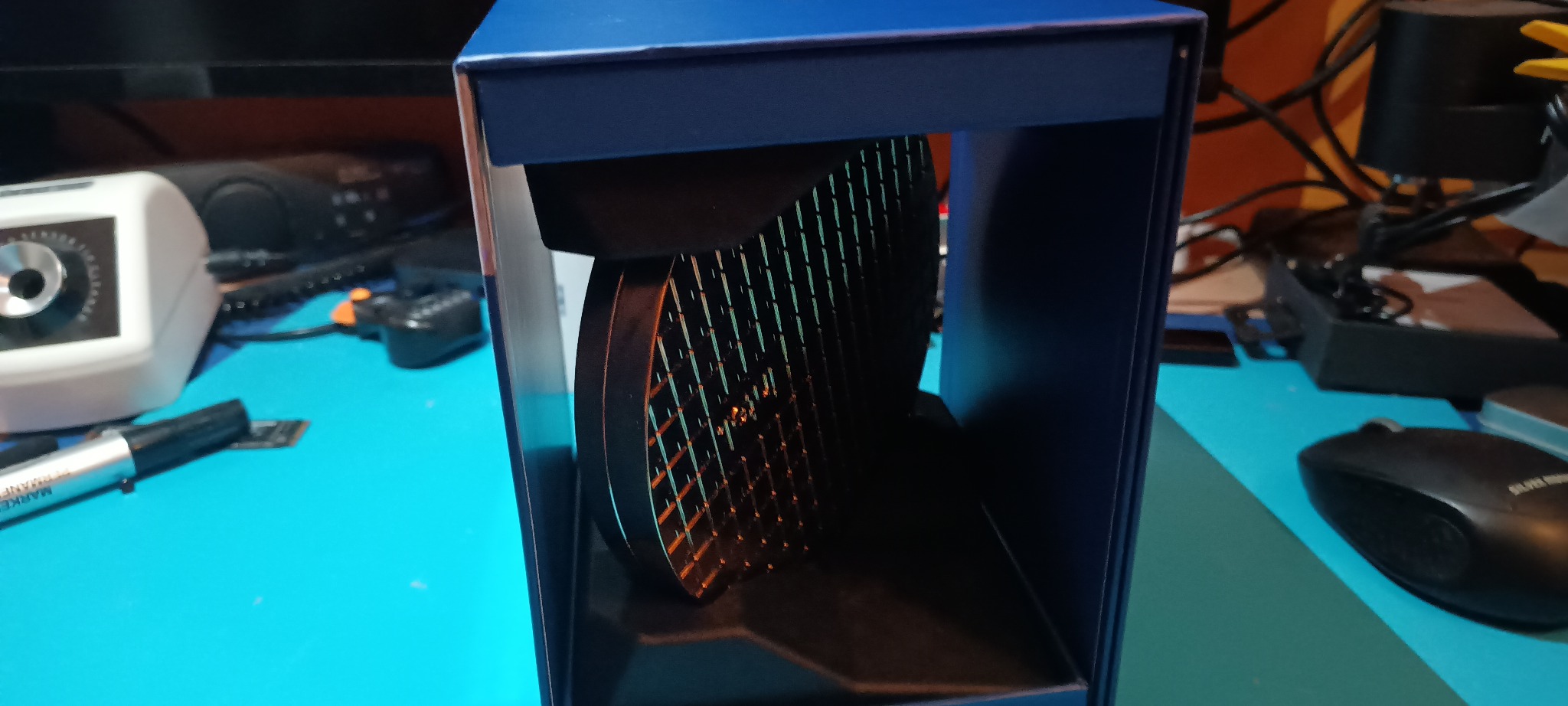 Dysk SSD w niebieskim pudełku na turkusowym biurku, obok mysz komputerowa i inne akcesoria. Widoczna struktura dysku z siatką. Ujęcie z bliska, oświetlenie studyjne.