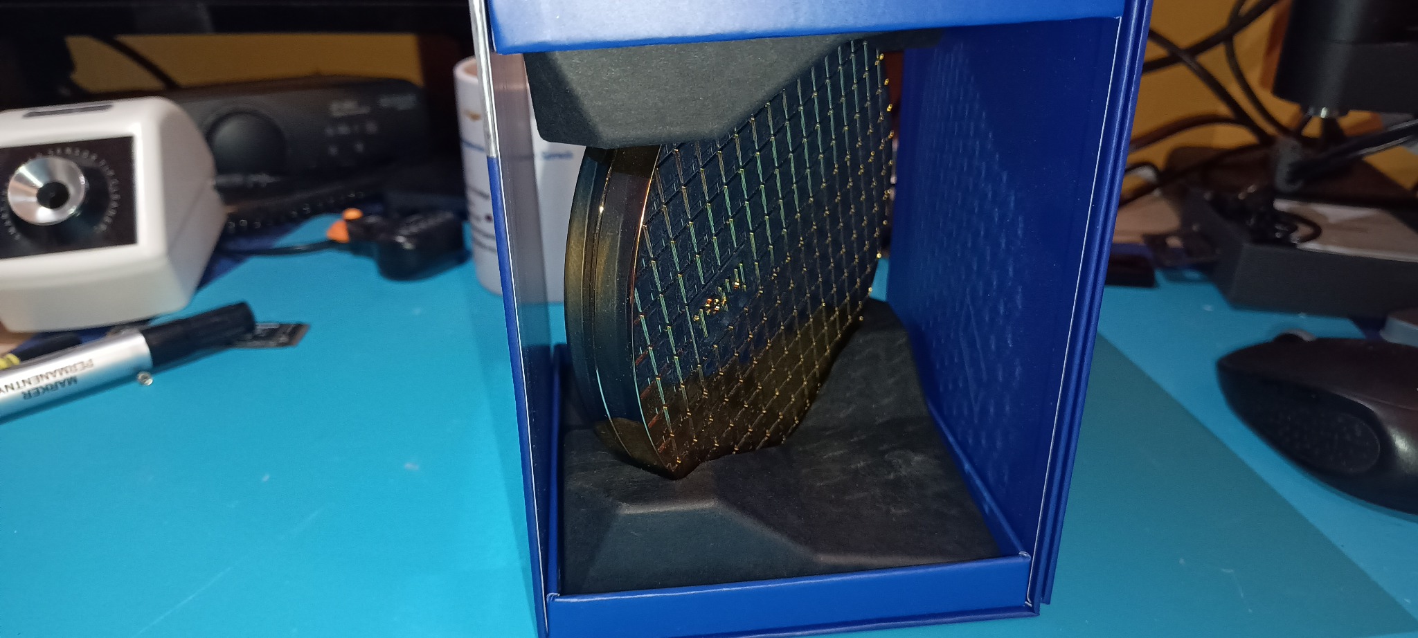Wafer krzemowy w niebieskim pudełku, widoczne struktury chipów. Na pierwszym planie marker permanentny i ołówek. Tło stanowią sprzęty elektroniczne i biurowe.