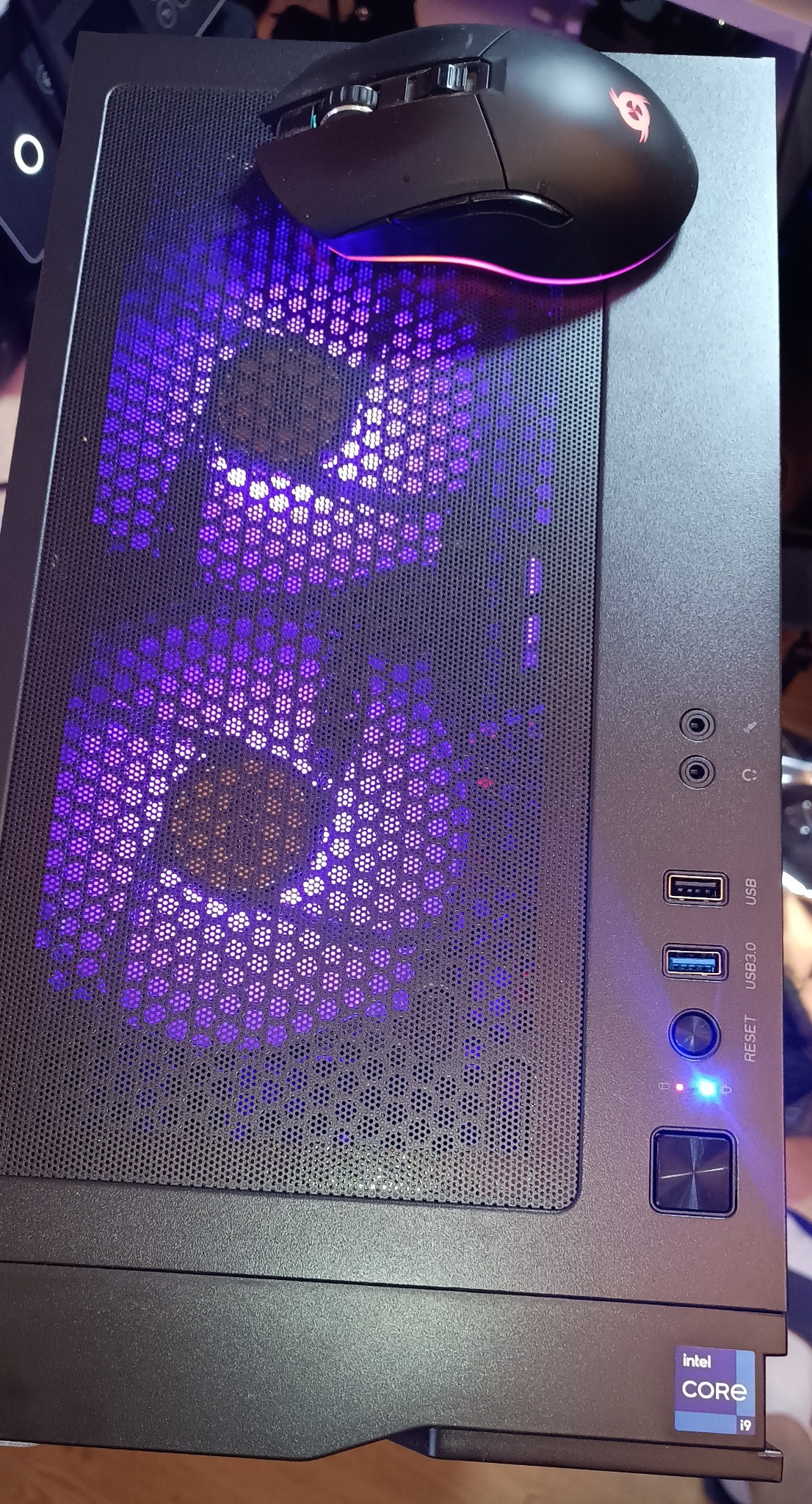 Czarna obudowa komputera stacjonarnego z podświetleniem LED w kolorze fioletowym, na której leży czarna myszka gamingowa z podświetleniem RGB.