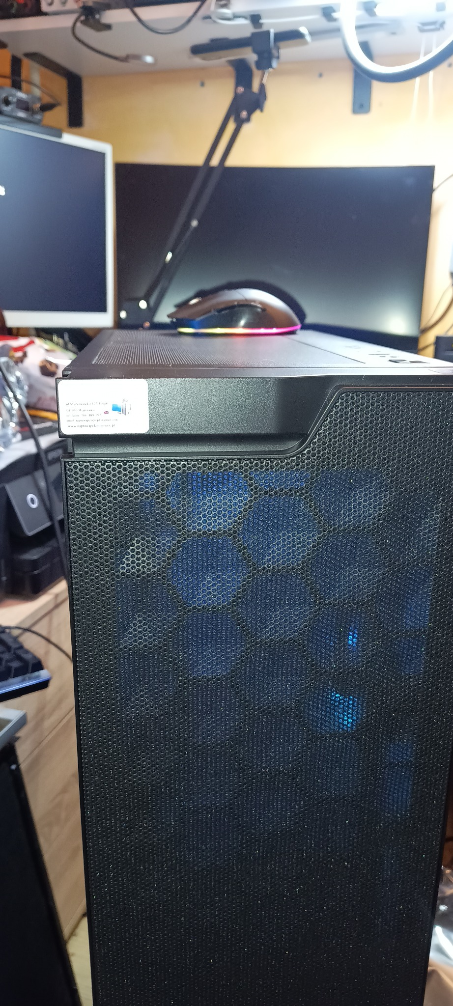 Czarna obudowa komputera stacjonarnego z widocznymi wentylatorami podświetlonymi na niebiesko, na górze myszka. Na obudowie naklejka z danymi firmy.