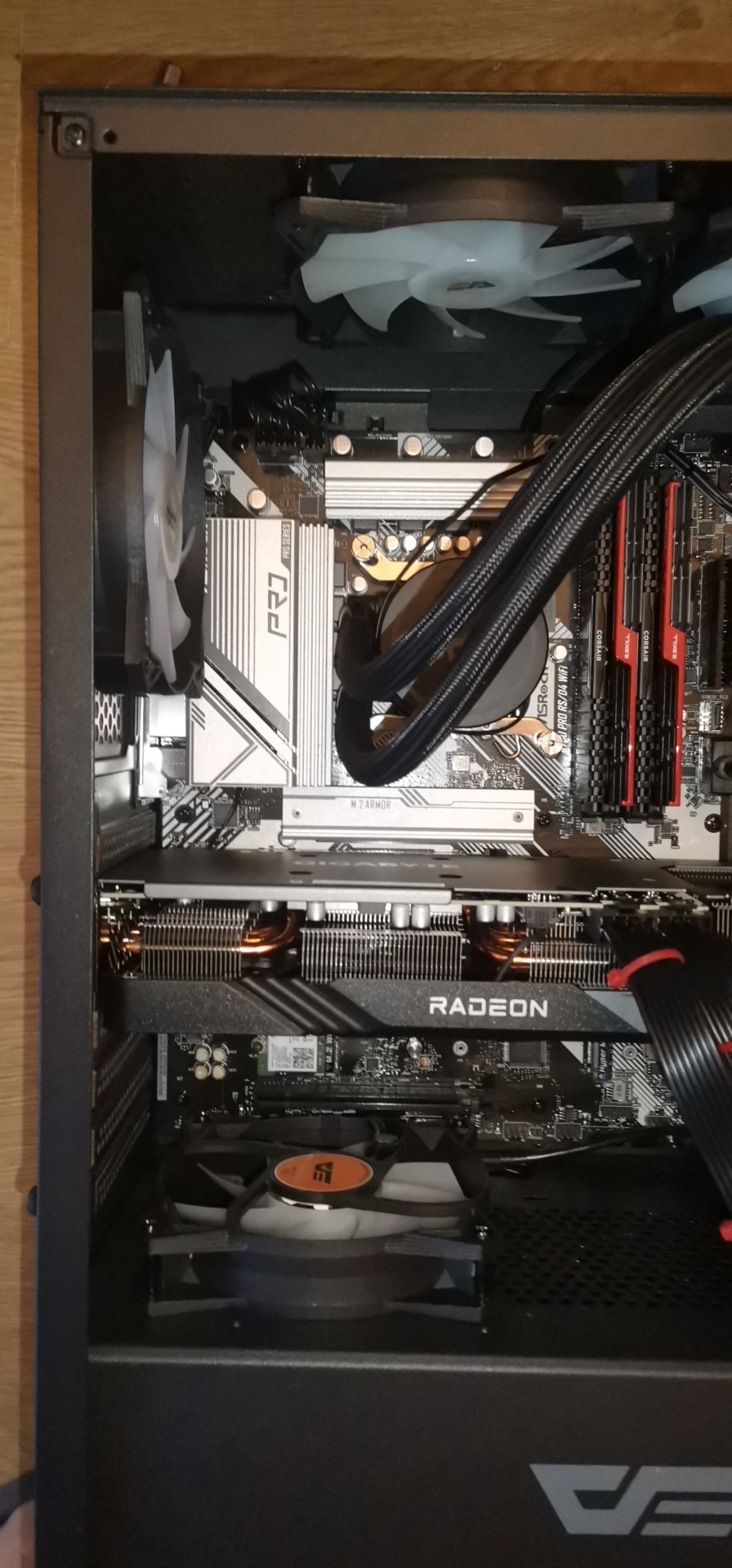 Wnętrze obudowy komputera stacjonarnego z widoczną kartą graficzną Radeon, płytą główną ASRock Pro Series, chłodzeniem wodnym i wentylatorami. Widoczne komponenty Corsair.