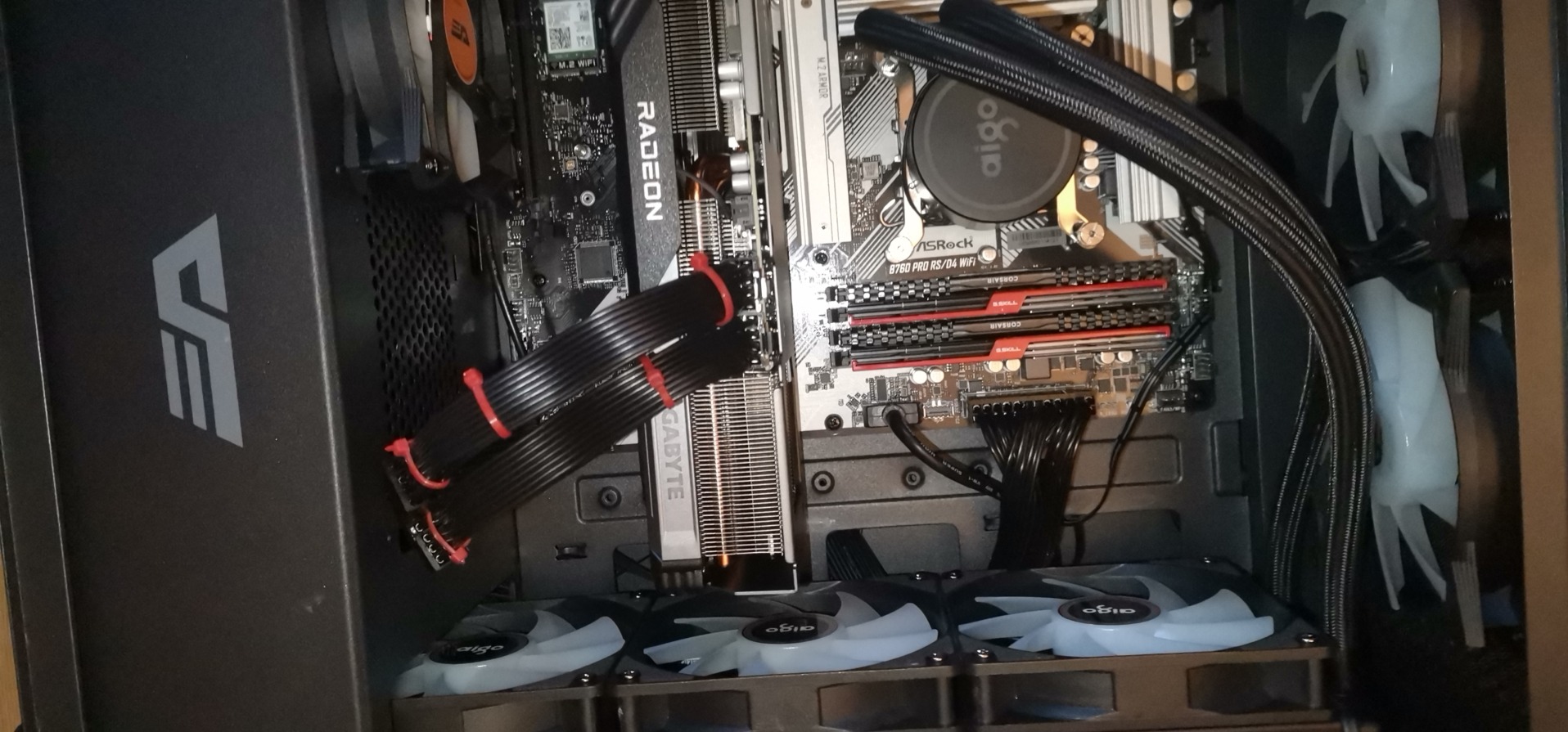 Wnętrze komputera stacjonarnego z widoczną płytą główną ASRock, kartą graficzną Radeon, pamięcią Corsair i chłodzeniem Aigo. Widoczne wentylatory i kable w obudowie.