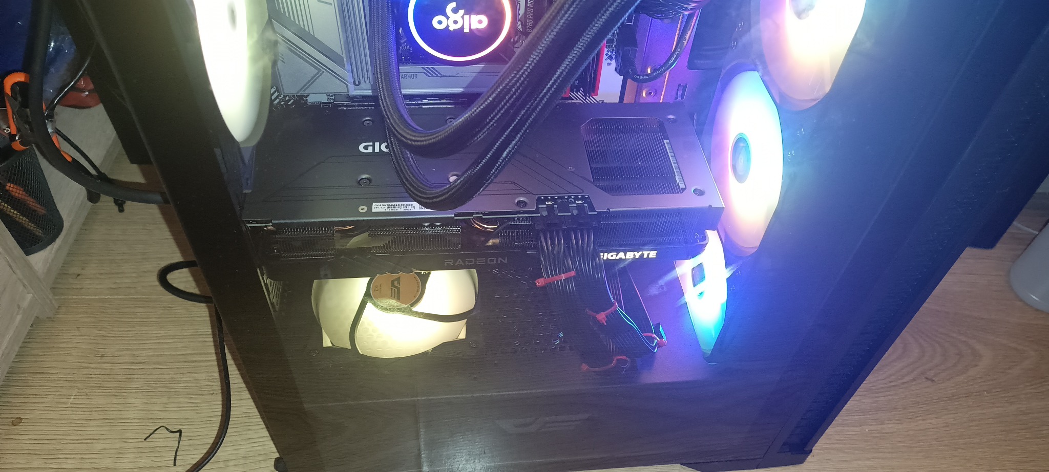 Wnętrze gamingowego PC z kartą graficzną Gigabyte Radeon, chłodzeniem wodnym Aigo i podświetlanymi wentylatorami. Widoczne kable i płyta główna. Całość w czarnej obudowie.