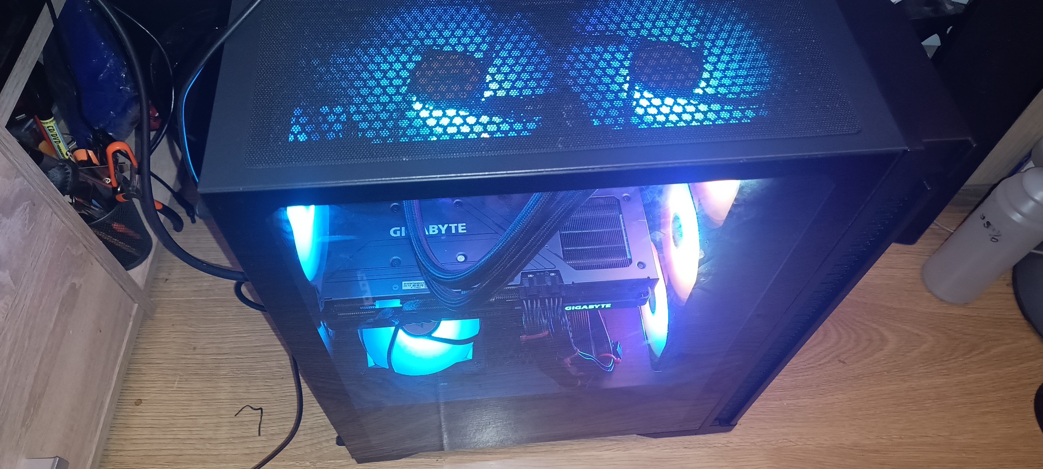 Komputer stacjonarny z podświetleniem LED w obudowie z oknem, widoczne komponenty Gigabyte, chłodzenie wodne. Ustawiony na podłodze, obok szafki i butelki.