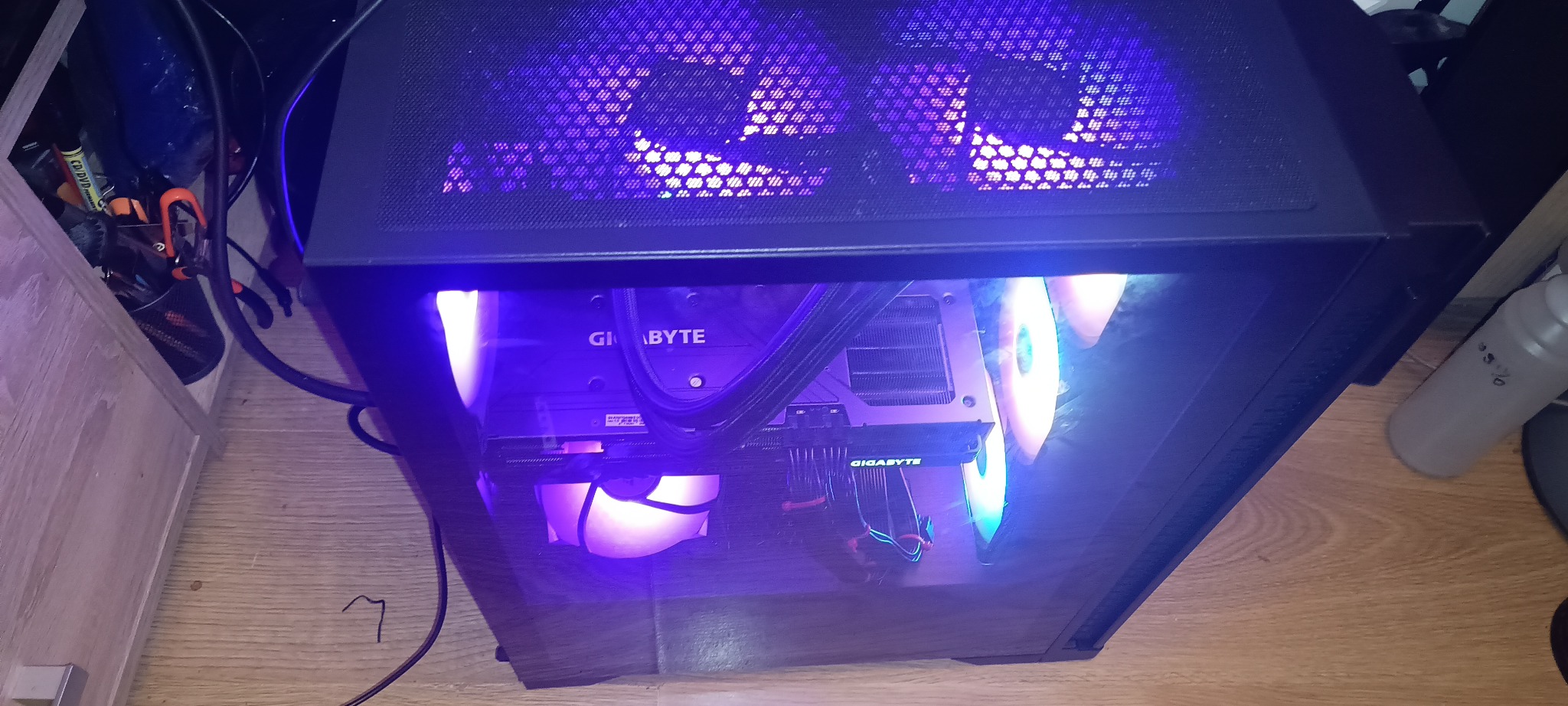 Czarna obudowa komputera stacjonarnego z widocznymi podświetlanymi wentylatorami i kartą graficzną Gigabyte, ustawiona na drewnianej podłodze.