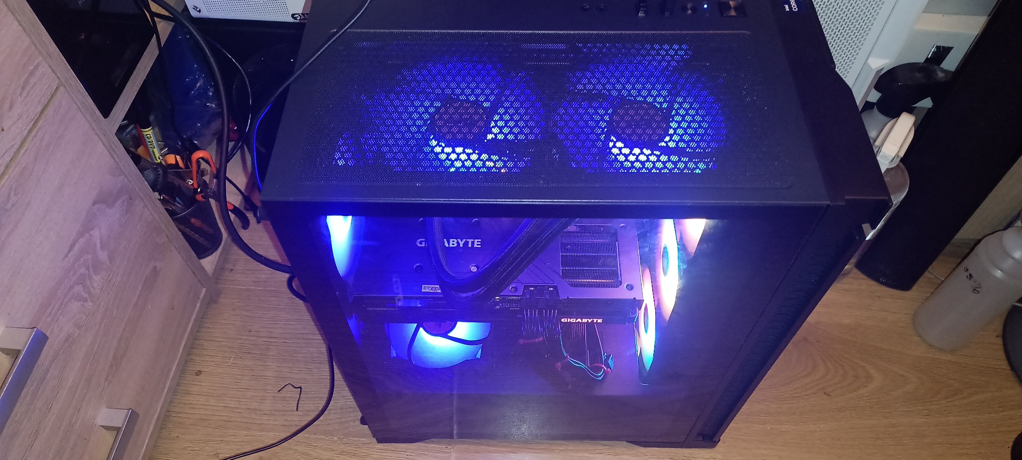 Komputer stacjonarny z podświetleniem LED w obudowie z oknem, widoczne komponenty marki Gigabyte. Obok szafka i butelka. Ujęcie z góry, lekko z boku.