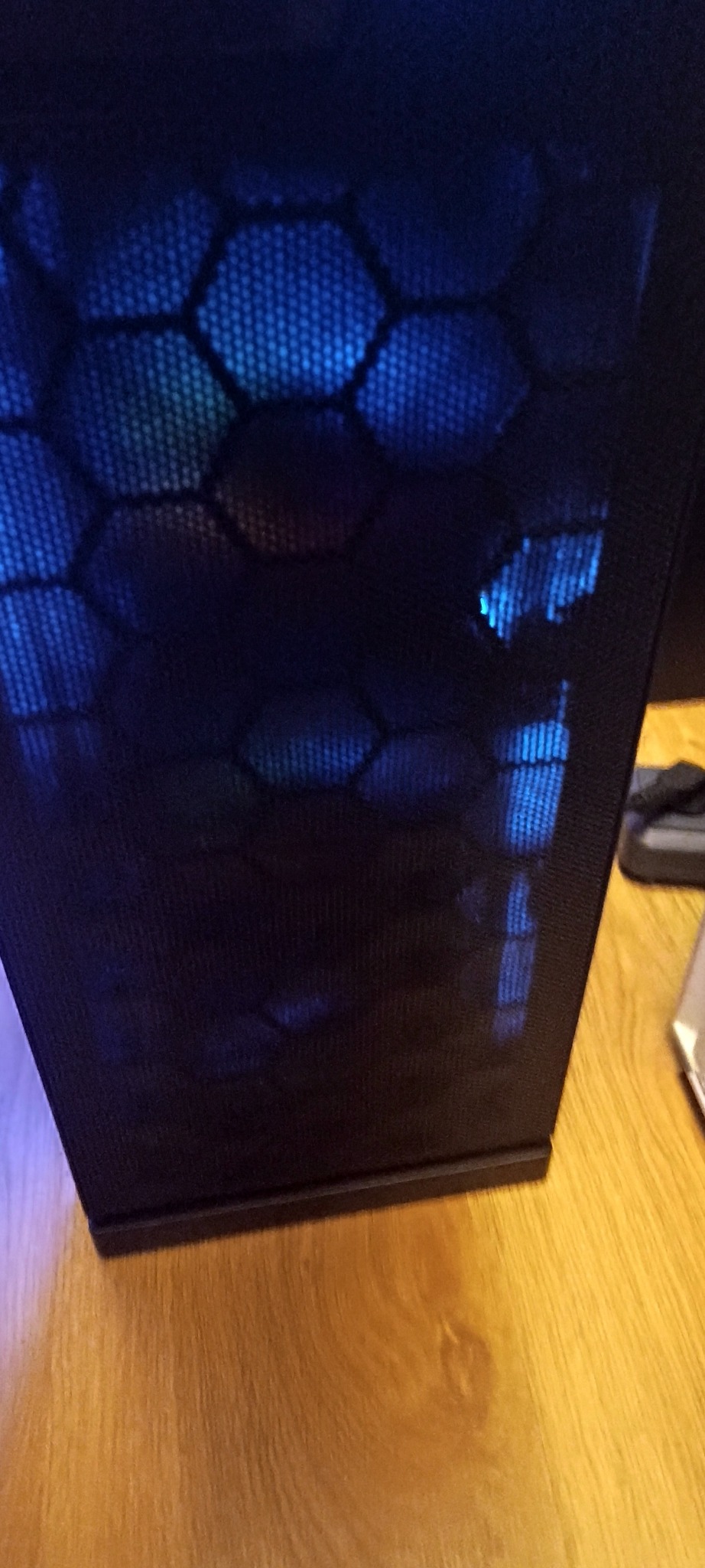 Częściowy widok obudowy komputera stacjonarnego z siatką wentylacyjną o strukturze plastra miodu, podświetloną niebieskim światłem LED. Widoczny fragment drewnianej podłogi.