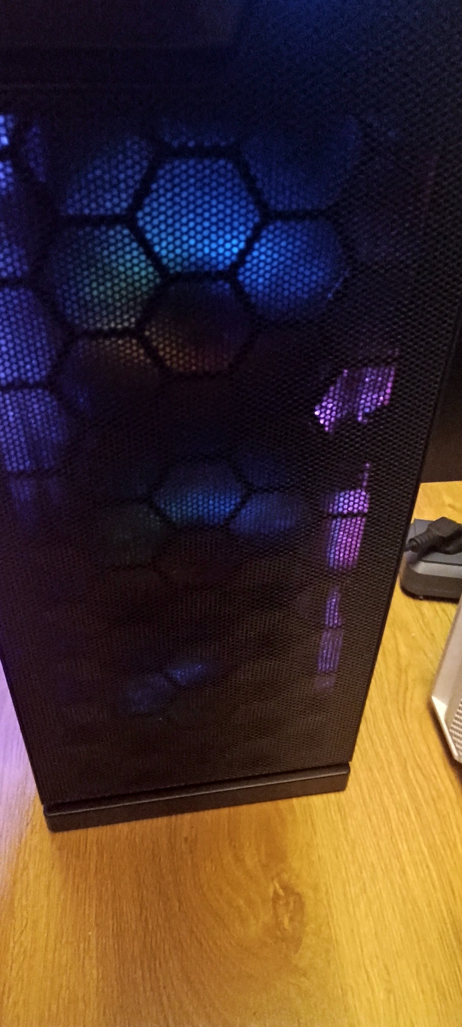 Czarna obudowa komputera stacjonarnego z siatką wentylacyjną i podświetleniem RGB w kształcie plastra miodu, stojąca na drewnianej podłodze.