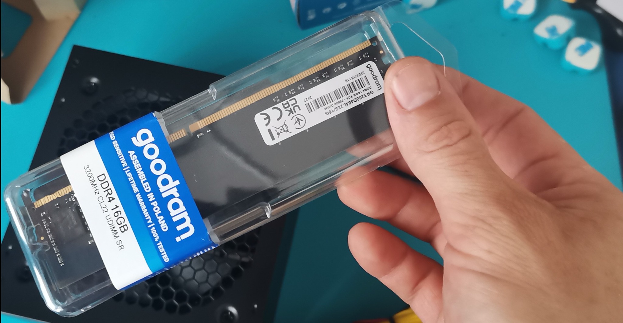 Pamięć RAM Goodram DDR4 16GB w przezroczystym opakowaniu, trzymana w dłoni nad czarną obudową komputera. Widoczna etykieta z informacjami technicznymi.
