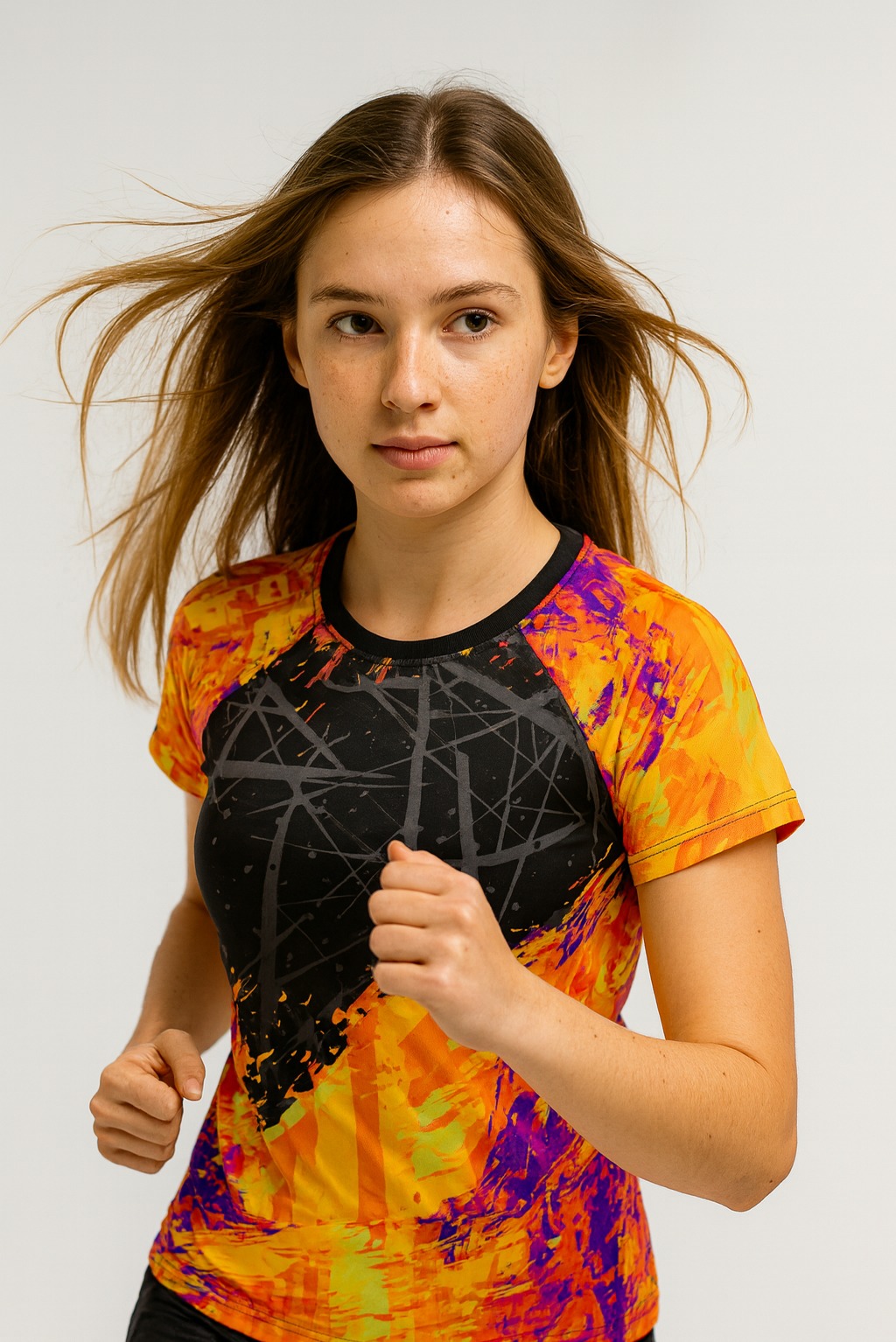 Kobieta w sportowej koszulce z abstrakcyjnym wzorem w odcieniach pomarańczy, fioletu i czerni, w dynamicznej pozie, na jasnym tle. Włosy w ruchu, skupiona twarz.