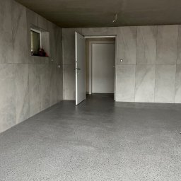 Life & Build - Ukończony garaż z szarą posadzką żywiczną z czarno-białymi flekami, ściany pokryte wielkoformatowymi płytkami w odcieniach szarości, otwarte białe drzwi.