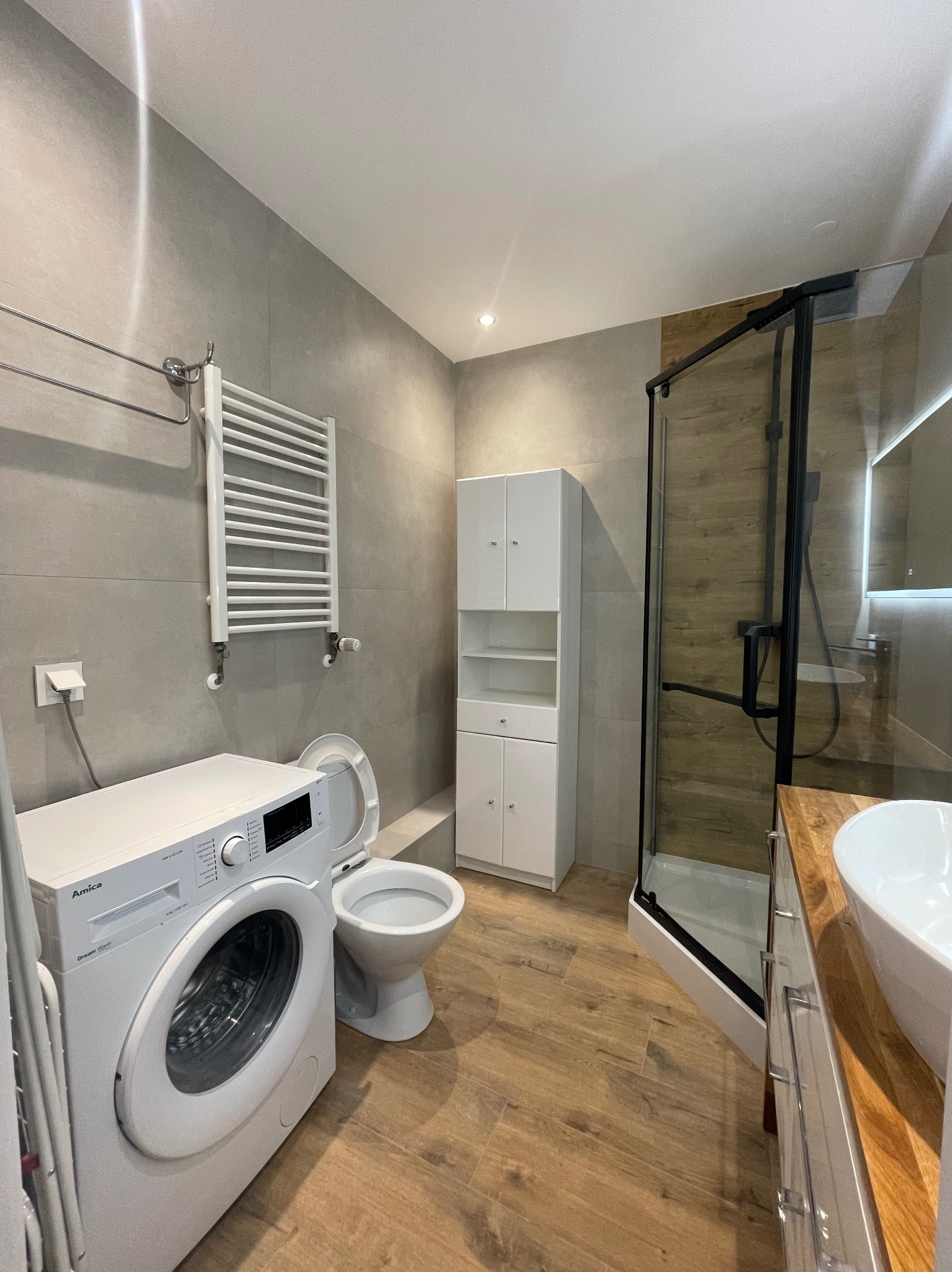 Szara łazienka po remoncie: pralka Amica, toaleta, kabina prysznicowa z czarną ramą, szafka i grzejnik drabinkowy. Płytki imitujące drewno na podłodze.