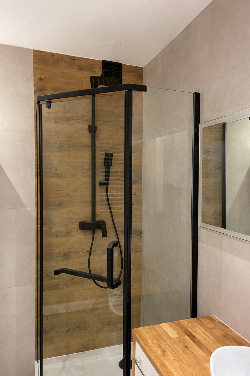 Nowoczesna kabina prysznicowa z czarnymi profilami i deszczownicą na tle ściany z drewnopodobnych płytek w jasnej łazience. Widoczny fragment blatu umywalki.