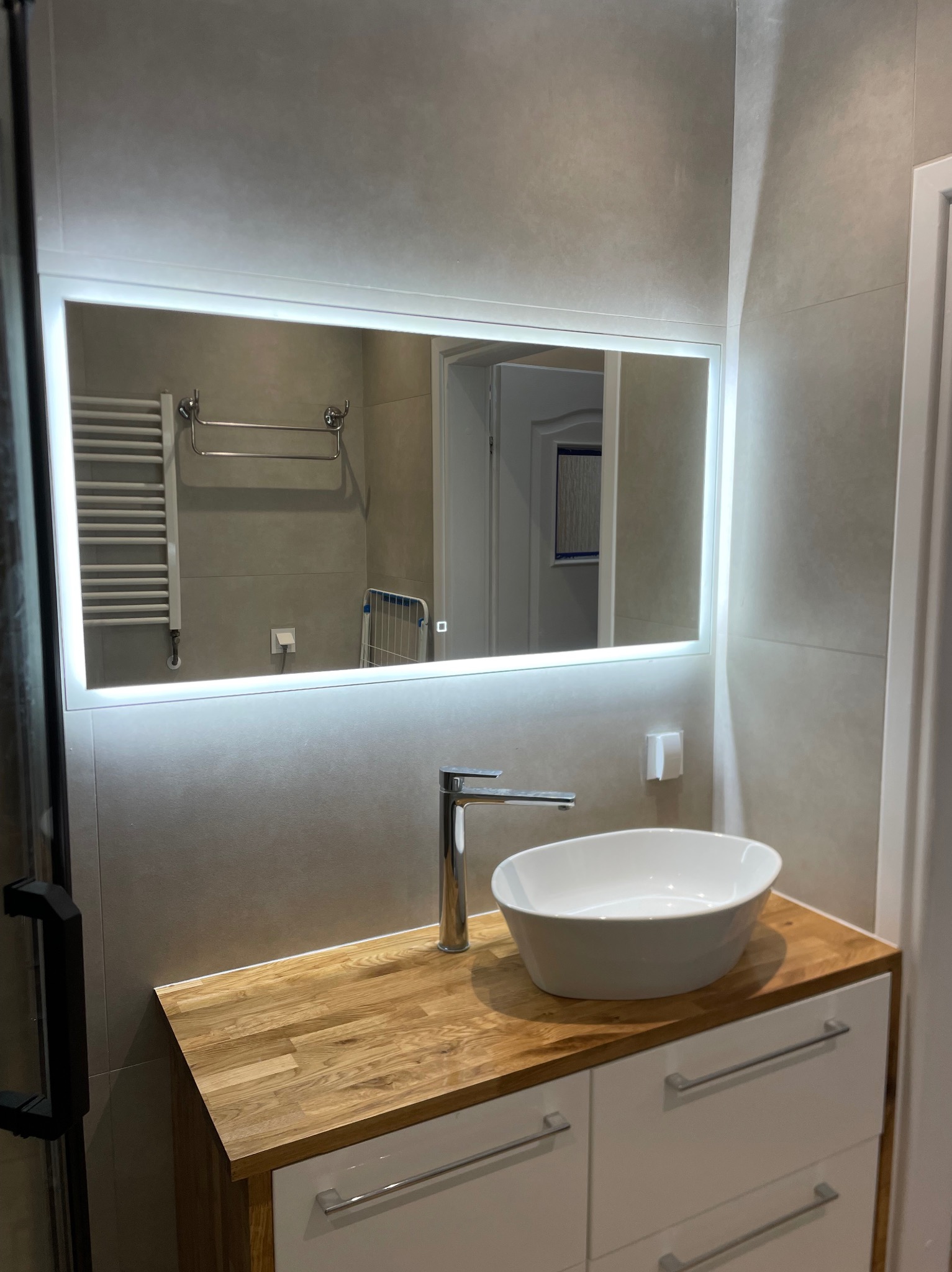 Nowoczesna łazienka z drewnianym blatem, białą umywalką nablatową i podświetlanym lustrem. W odbiciu widoczny grzejnik drabinkowy i suszarka na pranie.