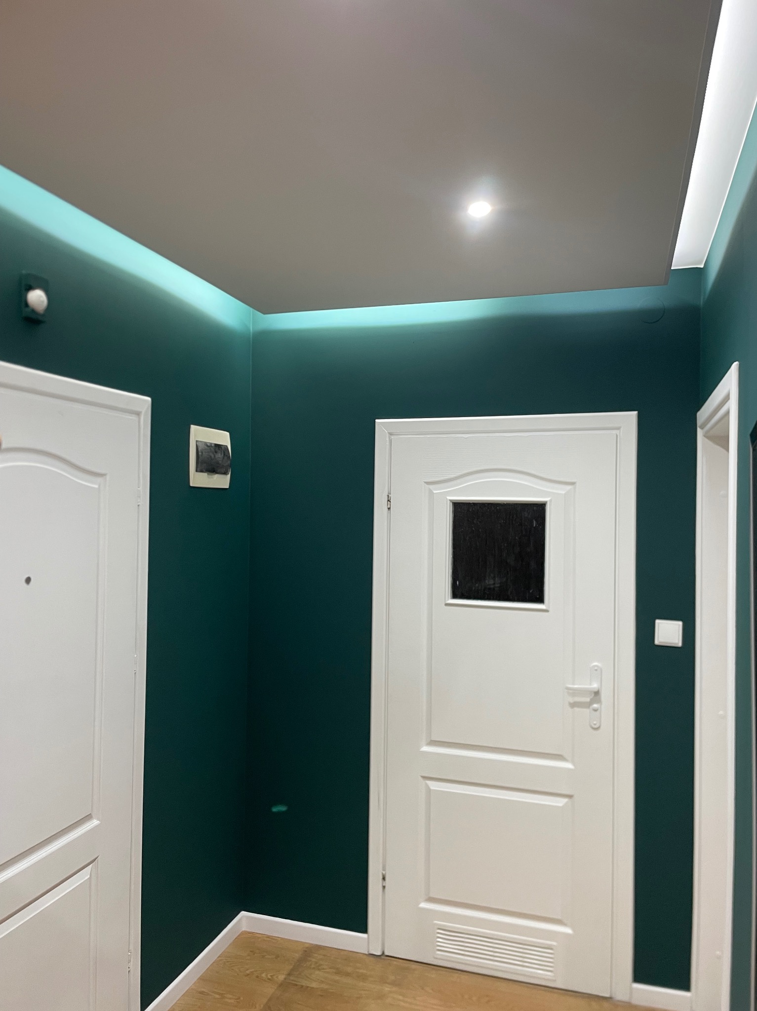 Korytarz po remoncie: ściany w odcieniach butelkowej zieleni, białe drzwi z szybką, podświetlany sufit. Widoczny fragment podłogi z paneli.