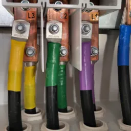 Podłączenie przewodów elektrycznych oznaczonych kolorami (żółty, zielony, fioletowy, niebieski) do miedzianych szyn w rozdzielnicy. Widoczne oznaczenia L1, L2, L3.