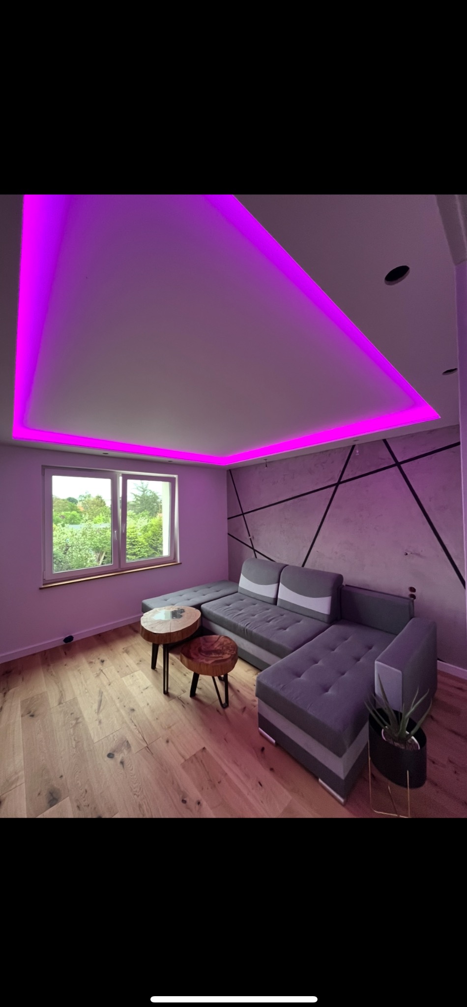 Nowoczesny salon z szarą sofą narożną, drewnianym parkietem i podświetlanym na fioletowo sufitem. Na ścianie dekoracyjny wzór z czarnych linii.
