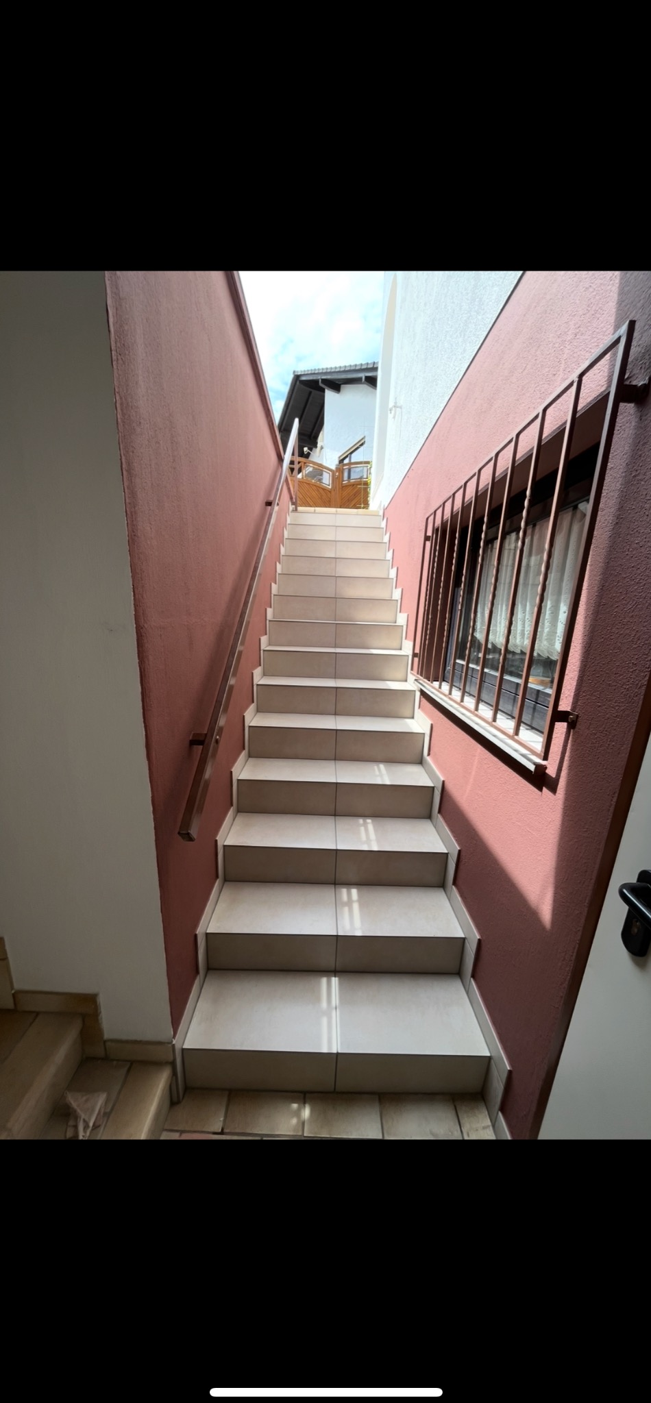 Wyłożone jasnymi płytkami schody zewnętrzne z metalową barierką i oknem z kratą, prowadzące do budynku. Ściany w kolorze terakoty i białe.