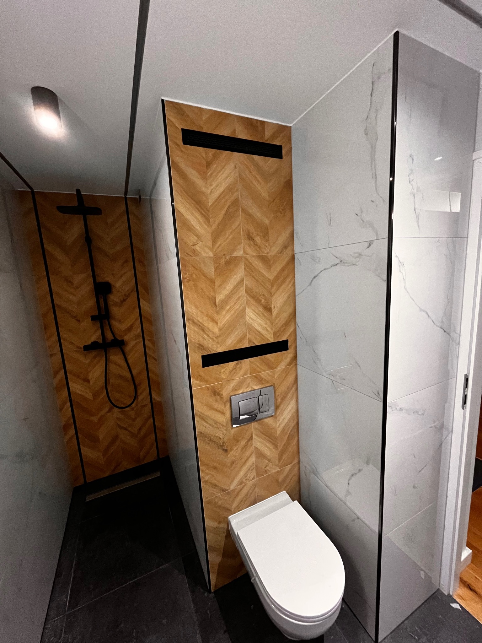 Nowoczesna łazienka z kabiną prysznicową, wykończona płytkami imitującymi drewno w jodełkę i białym marmurem. Wisząca toaleta i czarna armatura dodają elegancji.