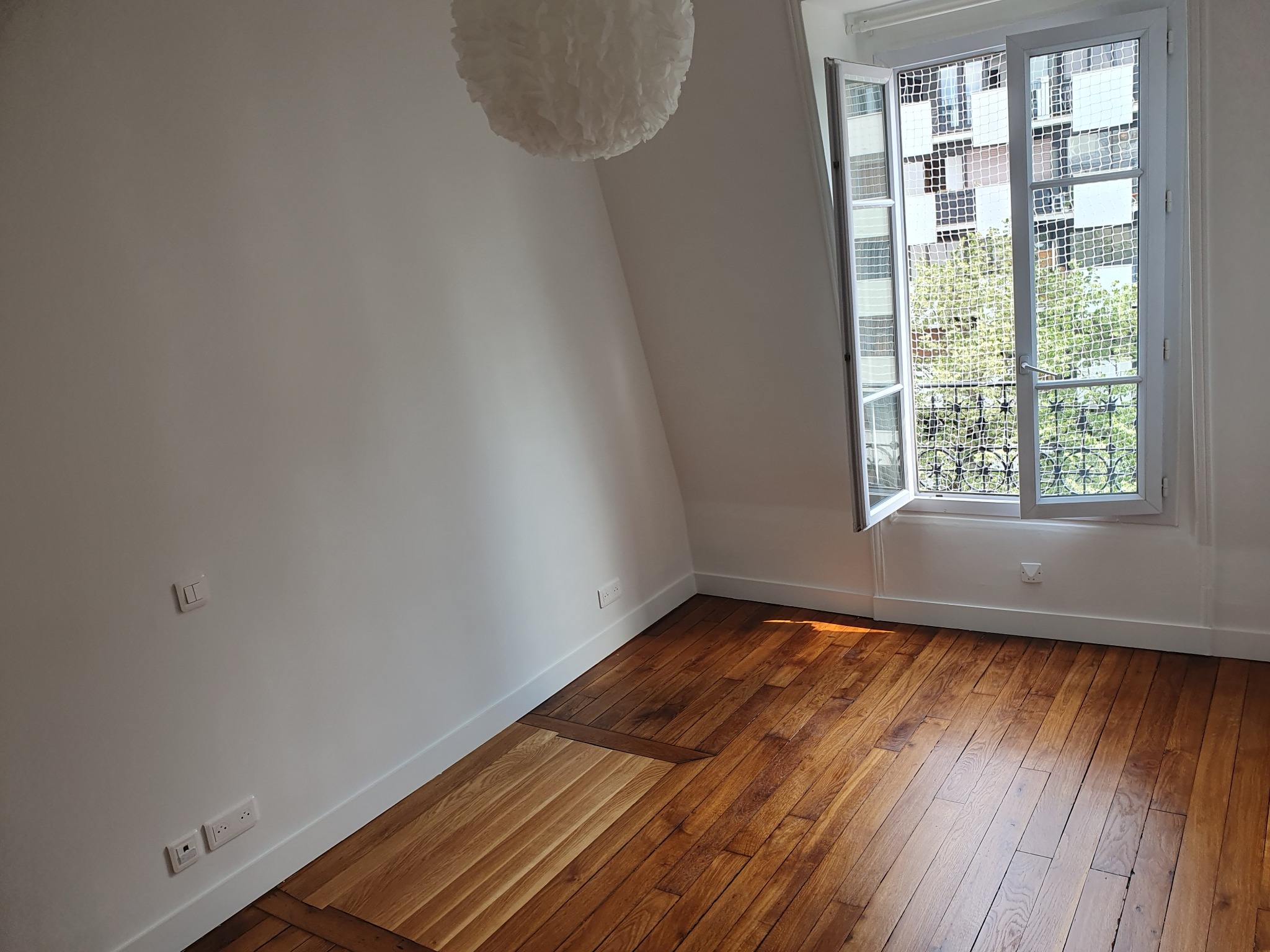 Jasne wnętrze poddasza z drewnianą podłogą i oknem z widokiem na zieleń. Białe ściany, dekoracyjna lampa. Widoczne gniazdka elektryczne przy podłodze.