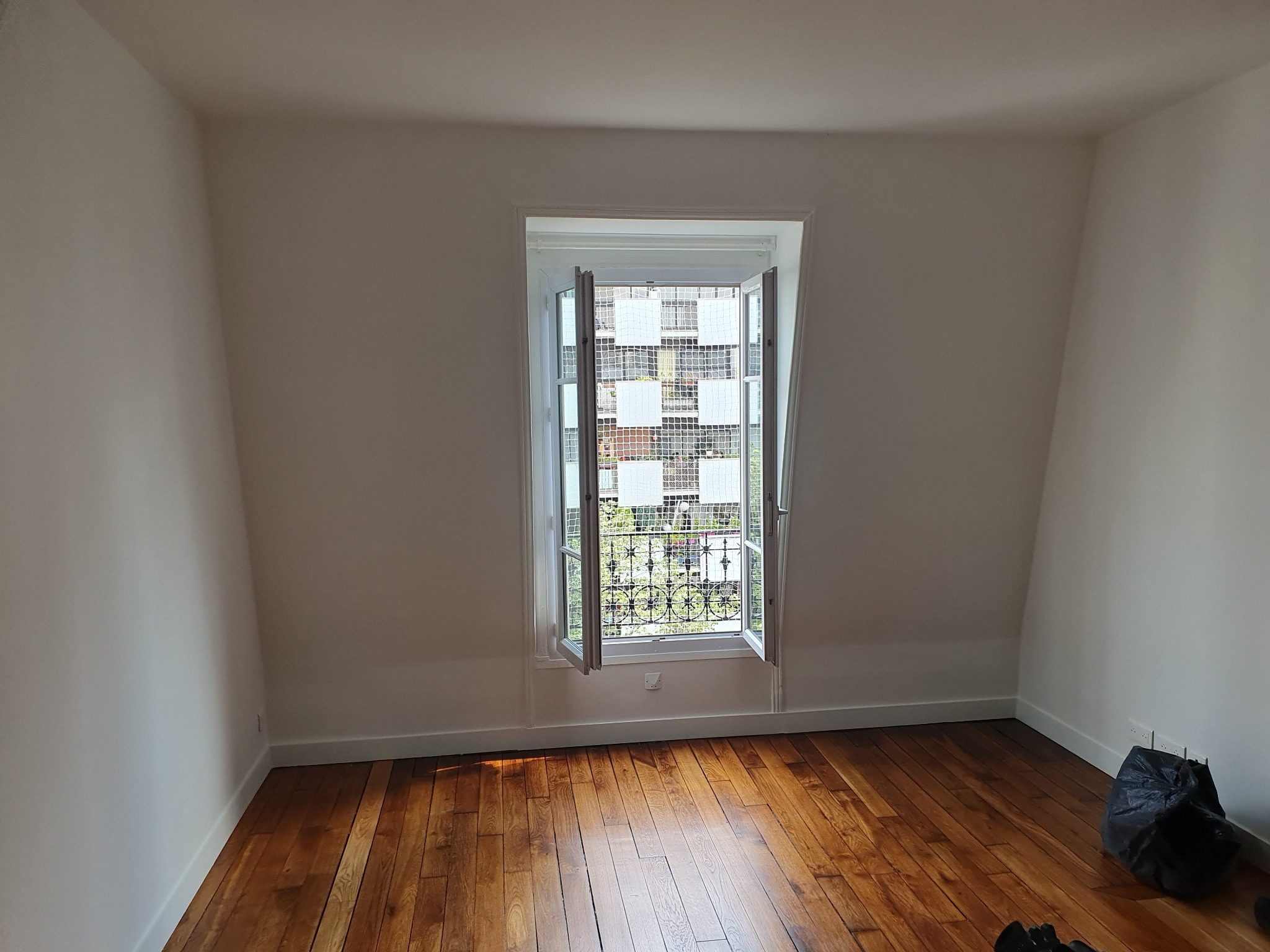 Jasne wnętrze poddasza z drewnianą podłogą i otwartym oknem. Świeżo pomalowane ściany w odcieniach bieli. W rogu widoczny worek na śmieci.