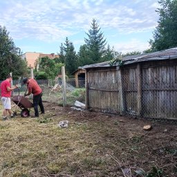 SZURU,BURU USŁUGI SPRZĄTAJĄCE DANIEL BARANOWSKI - Dwóch mężczyzn ładuje gałęzie do taczki na posesji z drewnianym budynkiem w tle. Widoczne ścięte pnie drzew i pozostałości po wycince. Praca w ogrodzie.