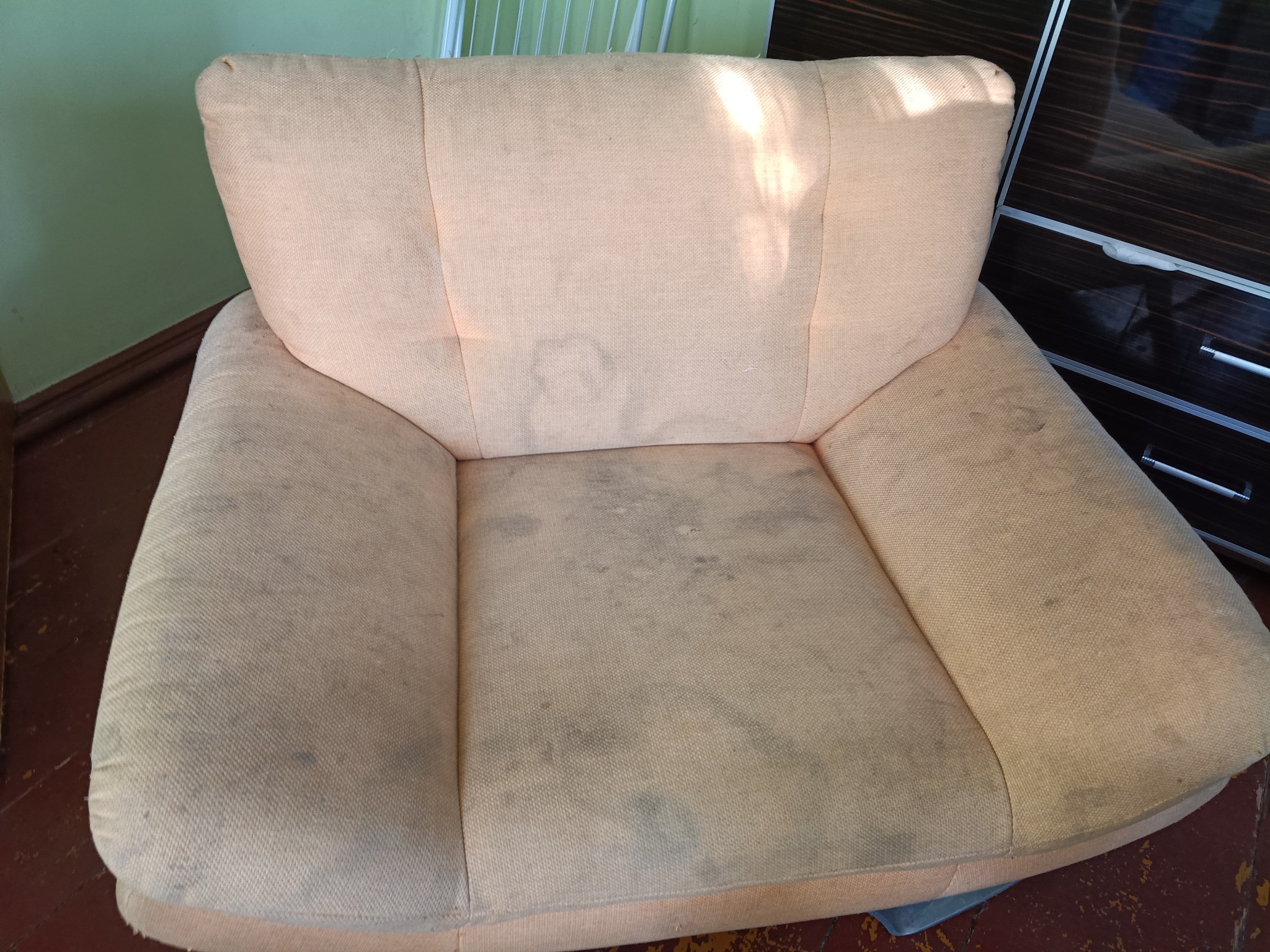 Zabrudzony, beżowy fotel tapicerowany z widocznymi plamami na siedzisku i oparciu, stojący na drewnianej podłodze, obok ciemnej szafy. Widoczne ślady użytkowania.