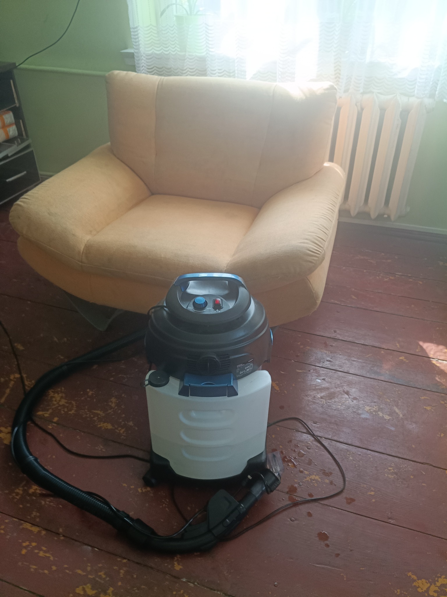 Profesjonalny odkurzacz piorący obok fotela w pokoju z drewnianą podłogą. Widoczne ślady wilgoci po praniu. Sprzęt gotowy do użycia, kabel zasilający podłączony.