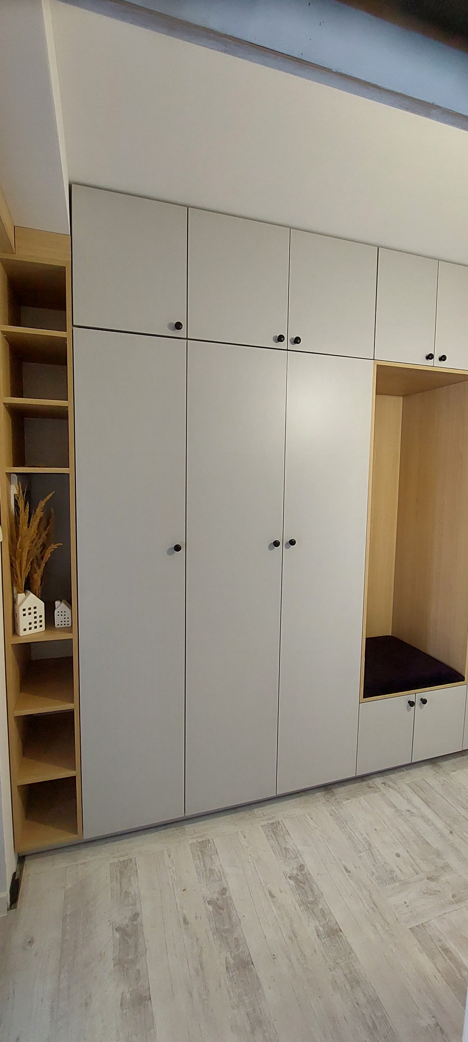 Jasnoszara szafa wnękowa z drewnianymi półkami i siedziskiem. Minimalistyczny design z czarnymi uchwytami. Dekoracje w postaci ceramicznych domków i suszonych traw.