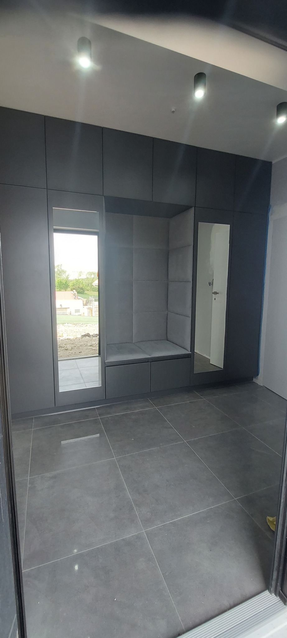 Szara szafa wnękowa z siedziskiem i lustrami, oświetlona punktowo, na tle szarej podłogi z płytek. Widok z perspektywy wejścia do pomieszczenia.