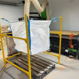 Mobilne Usługi Spawalnicze - Żółta metalowa konstrukcja wspierająca biały worek na odpady, obok maszyna CNC Pro 325 z systemem odsysania. Widoczne ślady obróbki drewna.