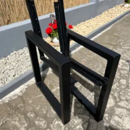 Surowe, czarne, metalowe ramy krzeseł na zewnątrz, z kwiatami w tle. Elementy konstrukcyjne krzeseł czekają na montaż siedzisk i oparć.