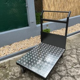 Mobilne Usługi Spawalnicze - Metalowy wózek transportowy z blachą ryflowaną, czarną ramą i czterema kołami, stojący na zewnątrz na tle zielonego ogrodzenia i bambusowej maty.