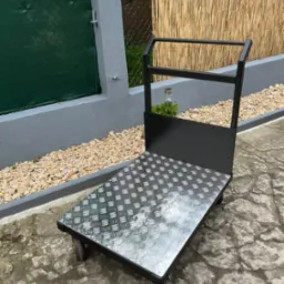 Metalowy wózek transportowy z blachą ryflowaną, czarną ramą i czterema kołami, stojący na zewnątrz na tle zielonego ogrodzenia i bambusowej maty.