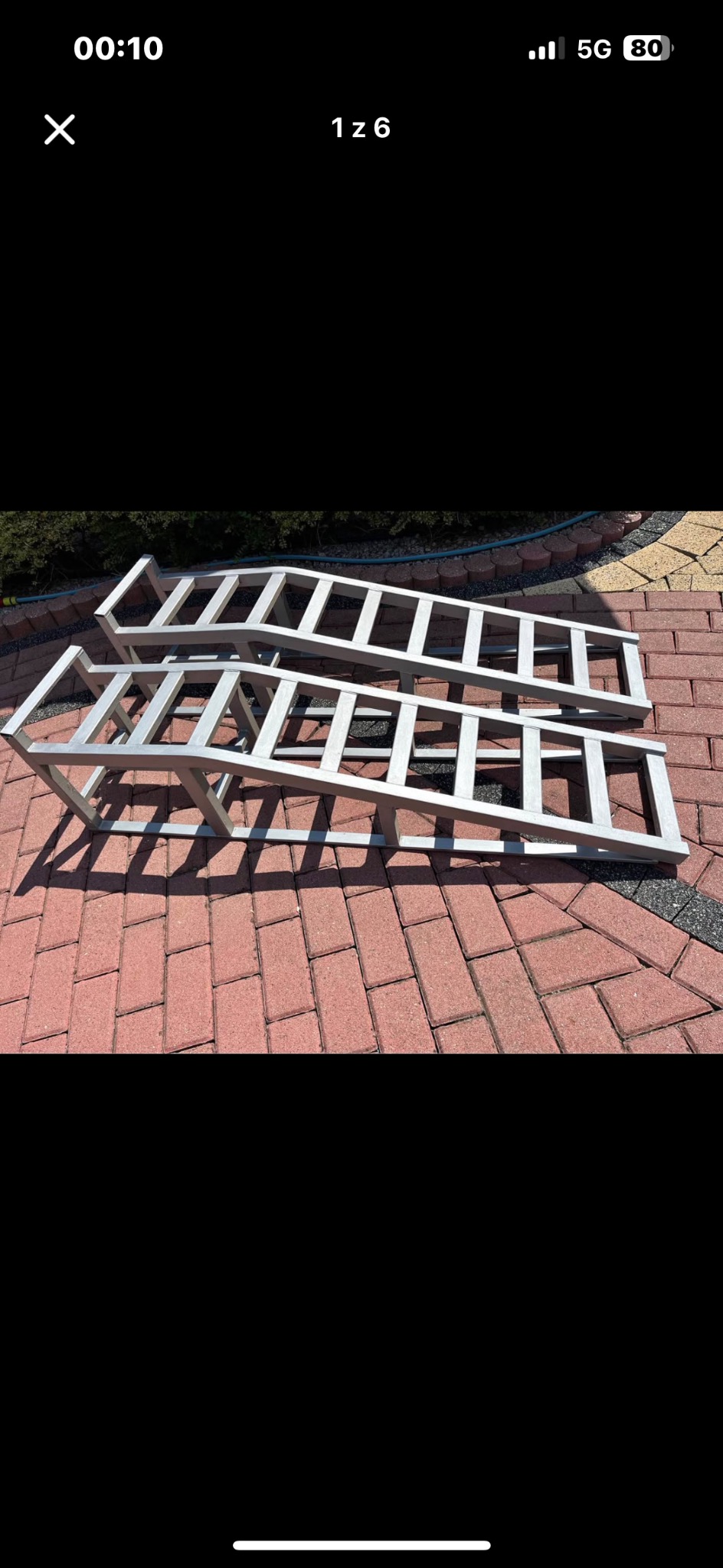 Dwie aluminiowe konstrukcje rampowe na czerwonej kostce brukowej, z zielenią w tle. Konstrukcje spawane, o regularnej strukturze, rzucają wyraźny cień.