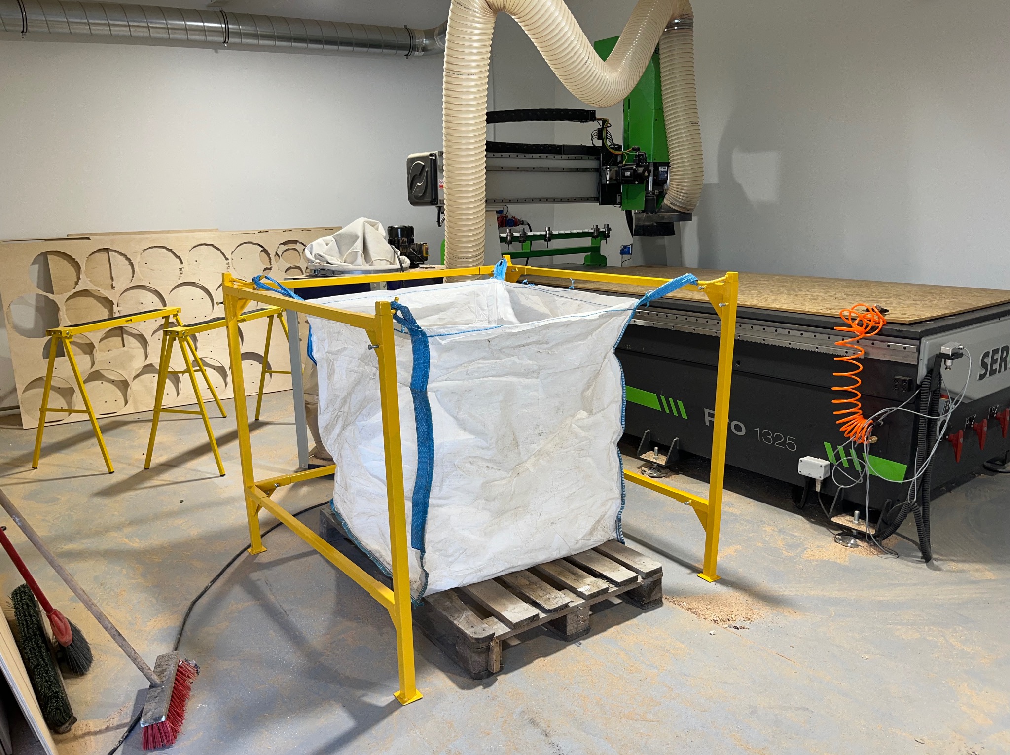 Wnętrze warsztatu z maszyną CNC, żółtą konstrukcją wsporczą i workiem na odpady. Widoczne narzędzia i elementy obróbki drewna. Jasne oświetlenie, surowy wystrój.