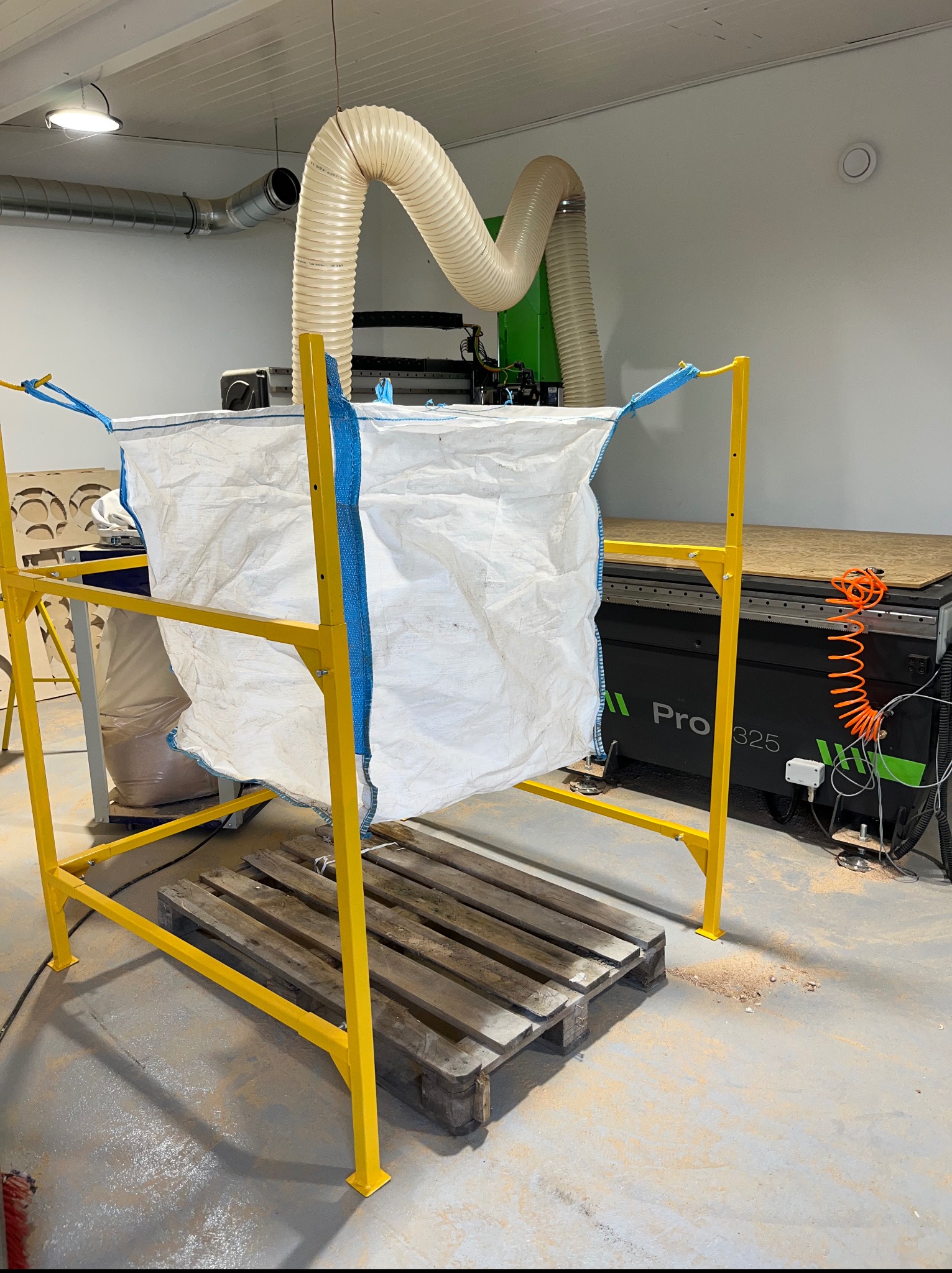 Żółta metalowa konstrukcja wspierająca biały worek na odpady, obok maszyna CNC Pro 325 z systemem odsysania. Widoczne ślady obróbki drewna.