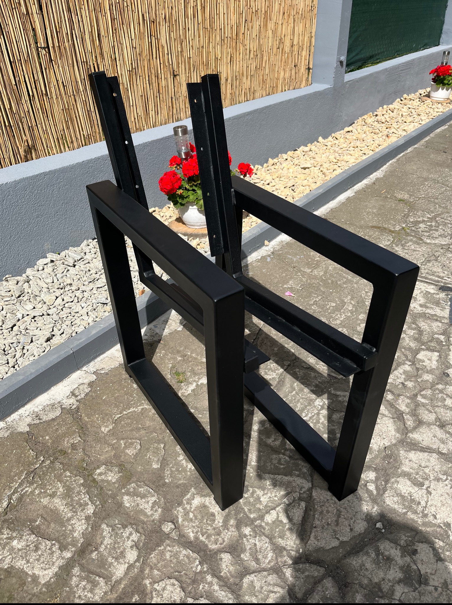 Surowe, czarne, metalowe ramy krzeseł na zewnątrz, z kwiatami w tle. Elementy konstrukcyjne krzeseł czekają na montaż siedzisk i oparć.