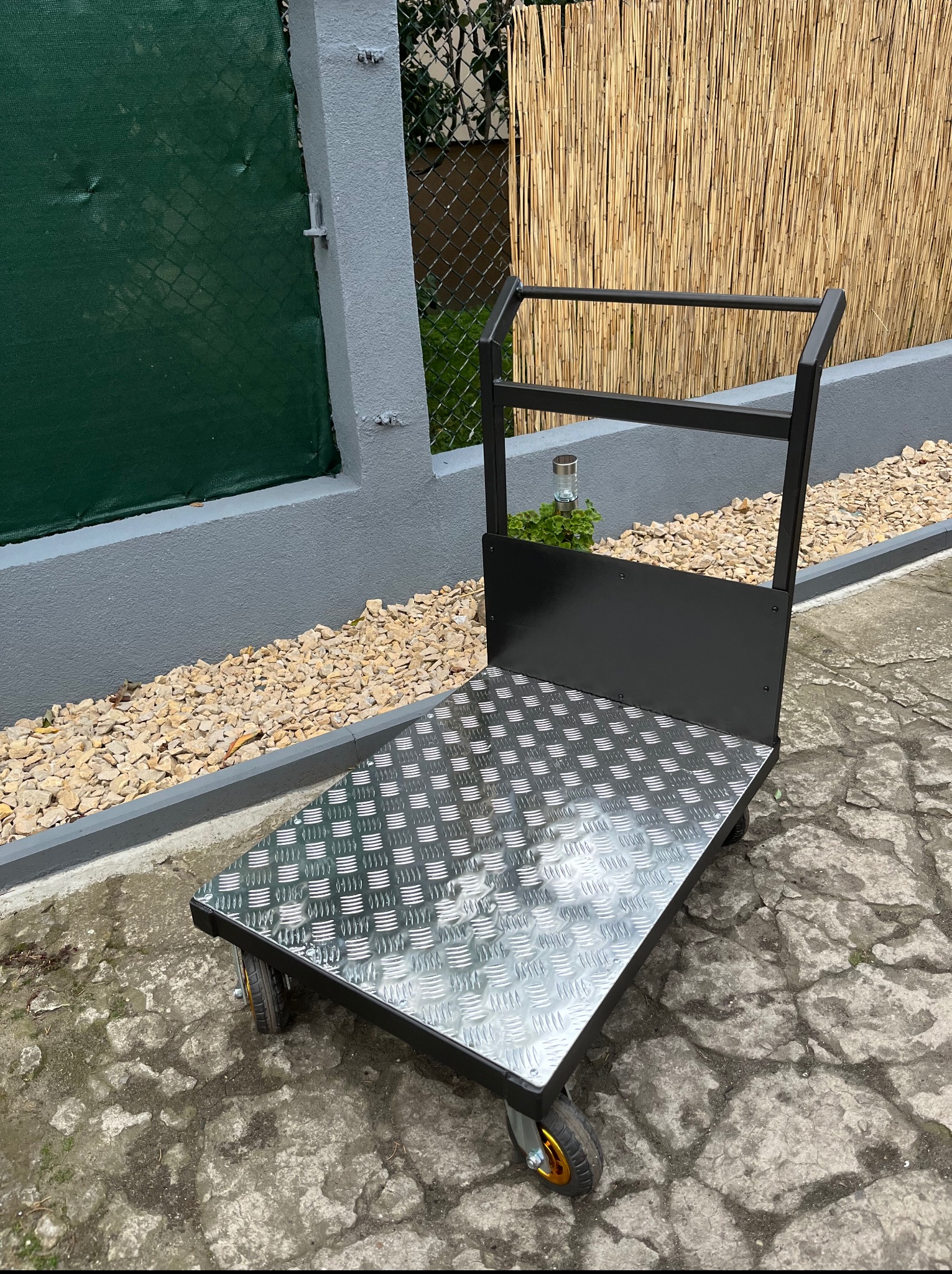 Metalowy wózek transportowy z blachą ryflowaną, czarną ramą i czterema kołami, stojący na zewnątrz na tle zielonego ogrodzenia i bambusowej maty.